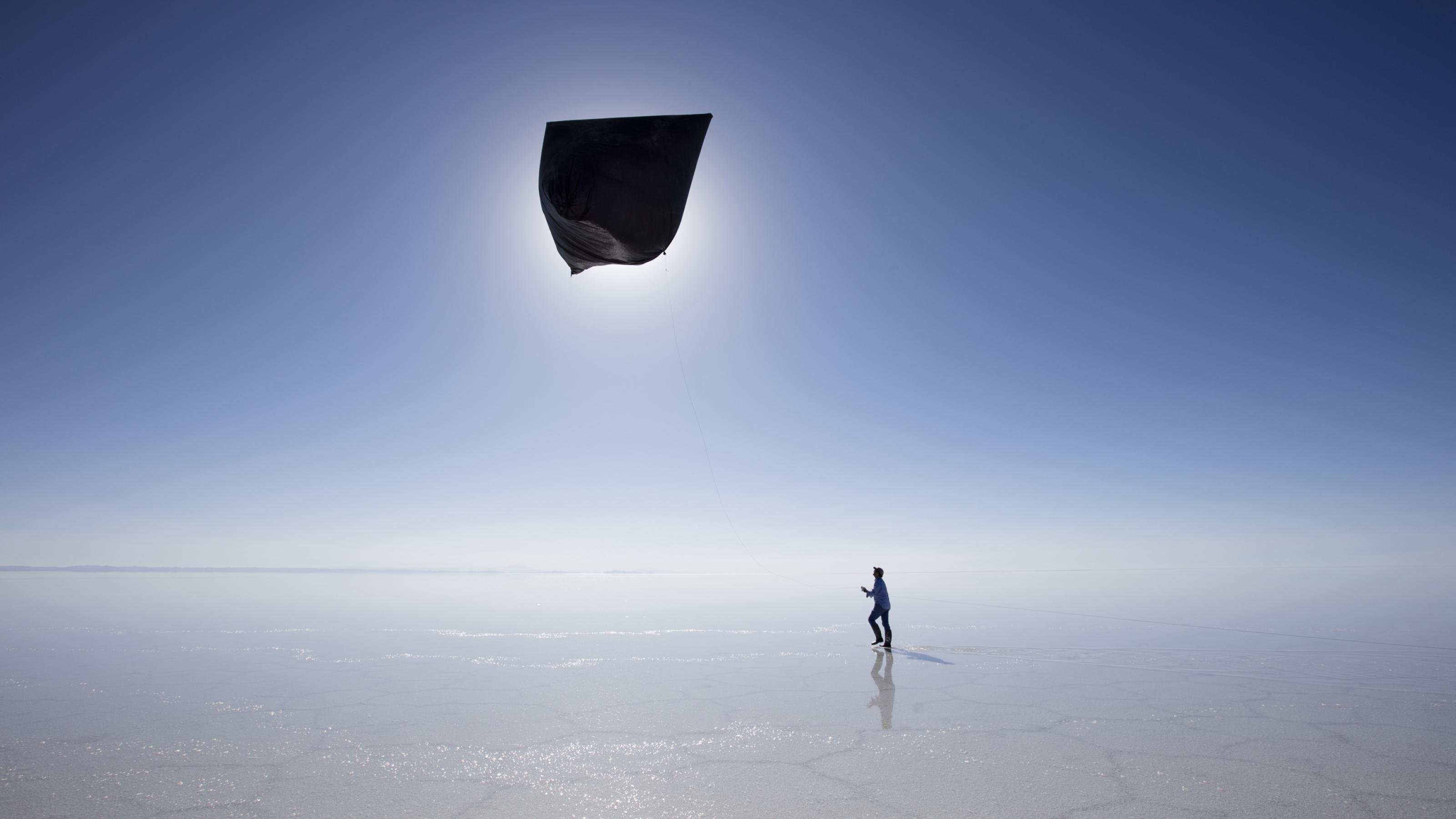 Vor blauem Himmel lässt ein Mann lässt in der Weite des Meeres bei Ebbe einen riesigen, schwarzen Drachen steigen.