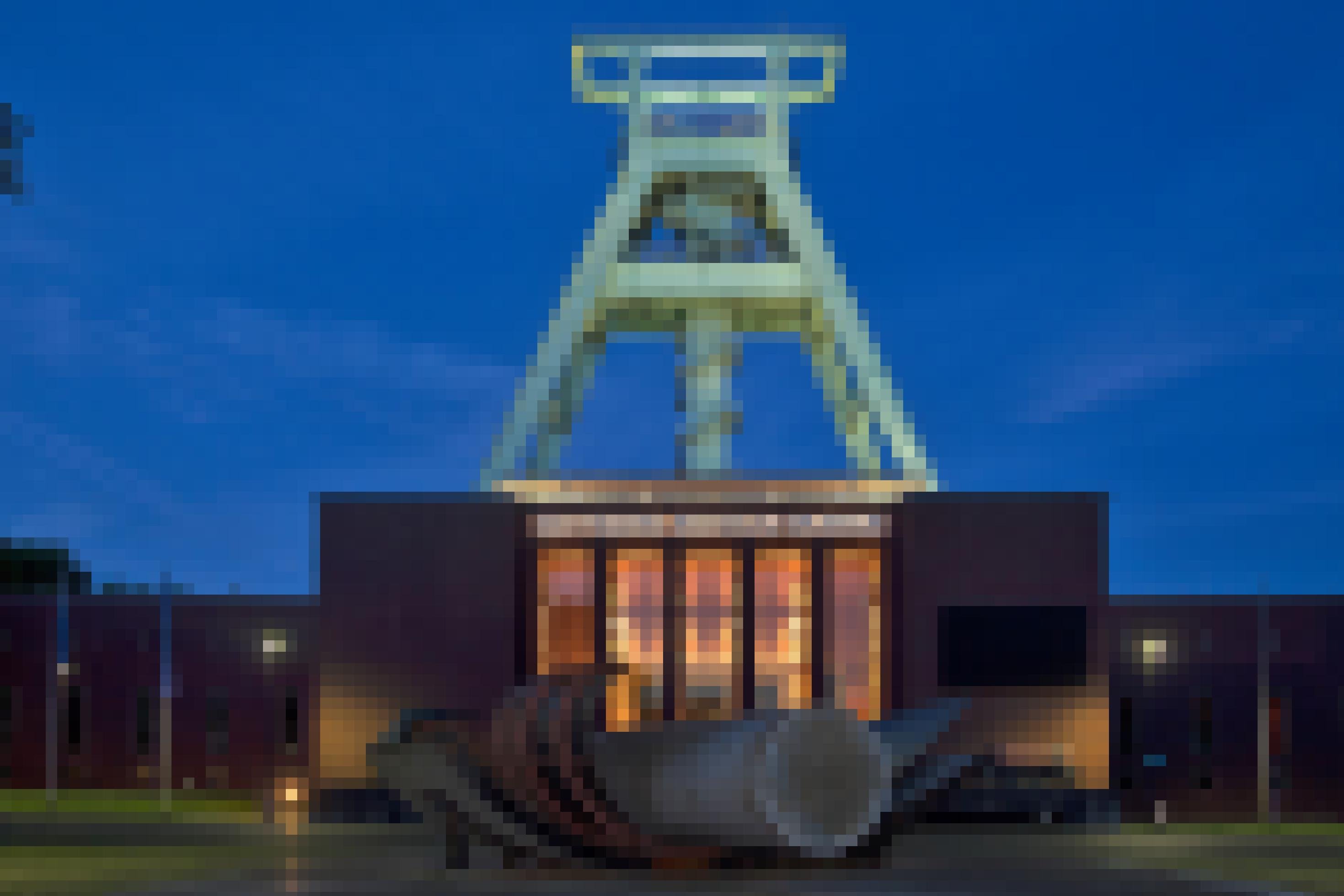 Das Foto zeigt ein beleuchtetes Fördergerüst hinter einem Gebäude, auf dem in Leuchtbuchstaben Deutsches Bergbau-Museum zu lesen ist.