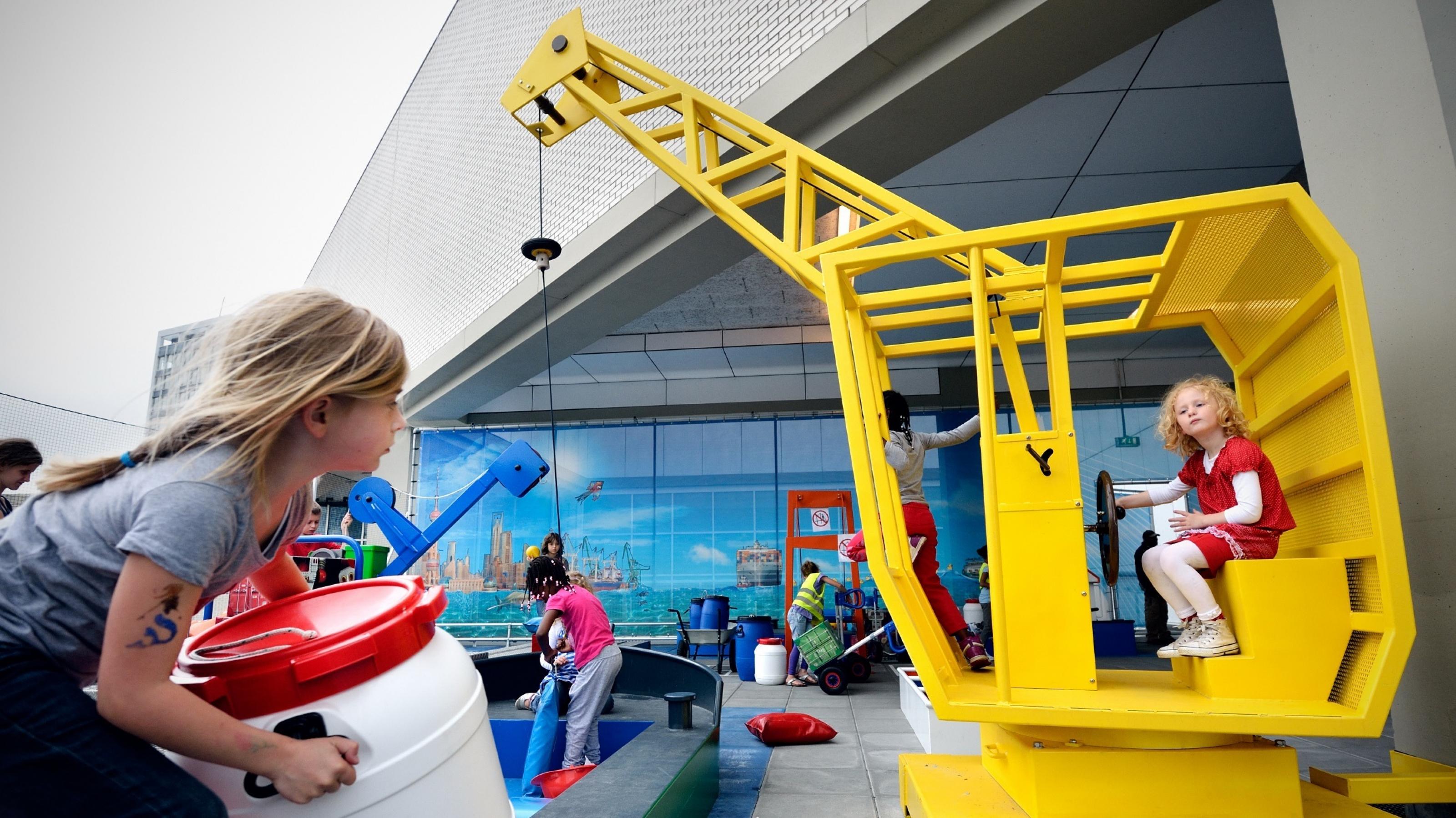 Das Foto zeigt ein Mädchen im Führerhaus eines Spielplatz-Krans, während ein anderes Kind ein Fass bereit stellt.