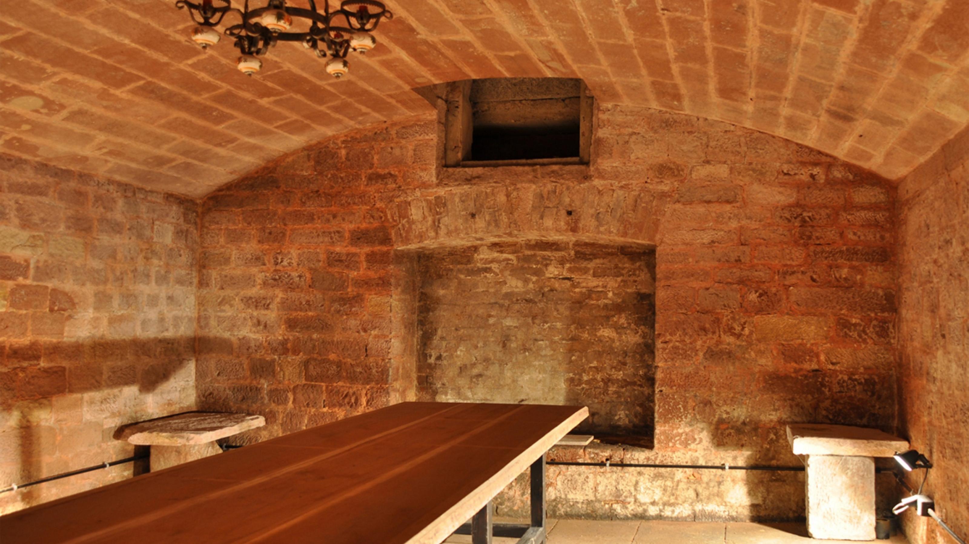 Das Foto zeigt die unverputzte Ziegelwand eines Gewölbekellers, einen langen Tisch, Bänke und einen von der Decke herabhängenden Leuchter.