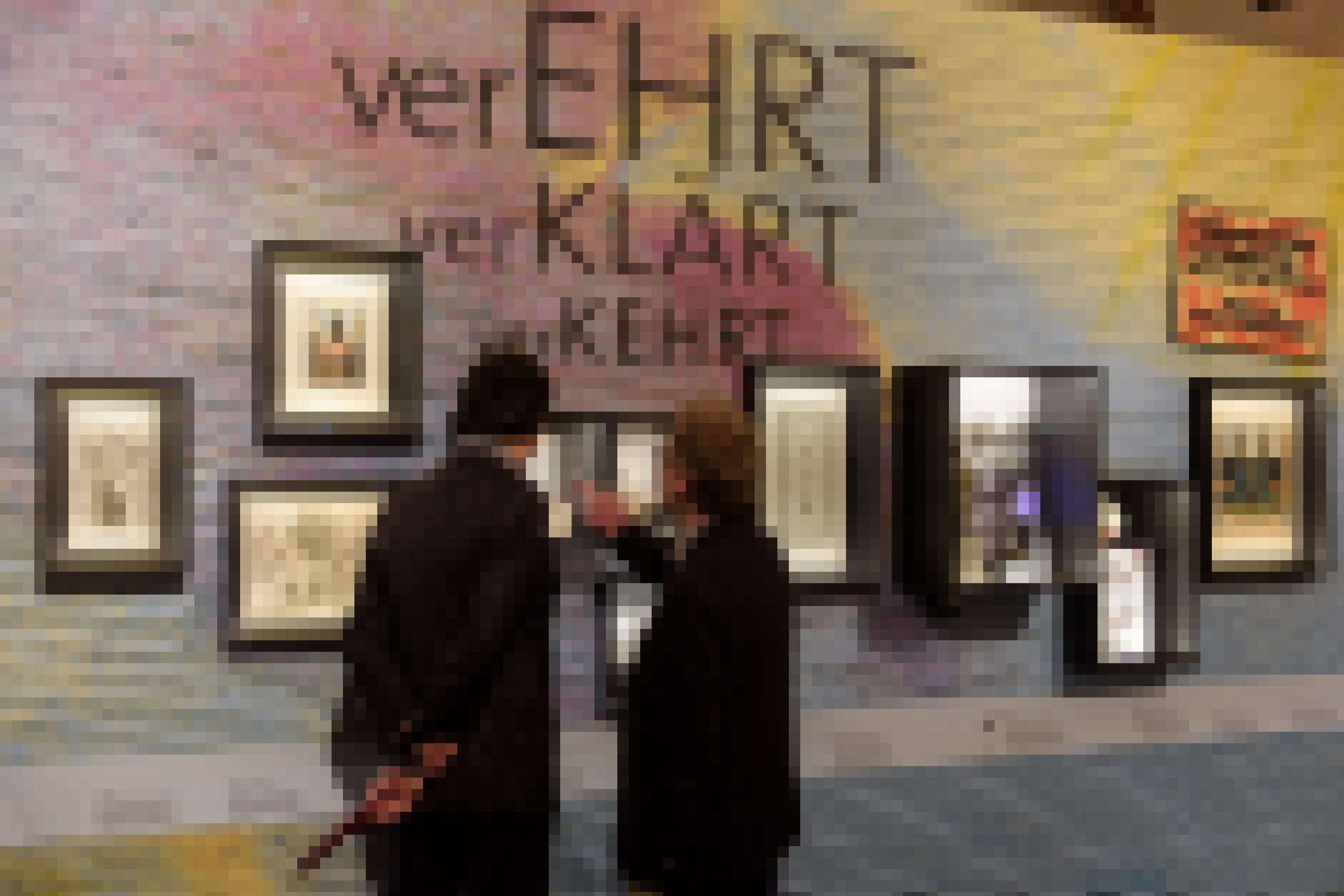 Zwei Männer stehen vor einer Wand, auf die – in unterschiedlicher Höhe – einzelne gerahmte Ausstellungsexponate gehängt sind.