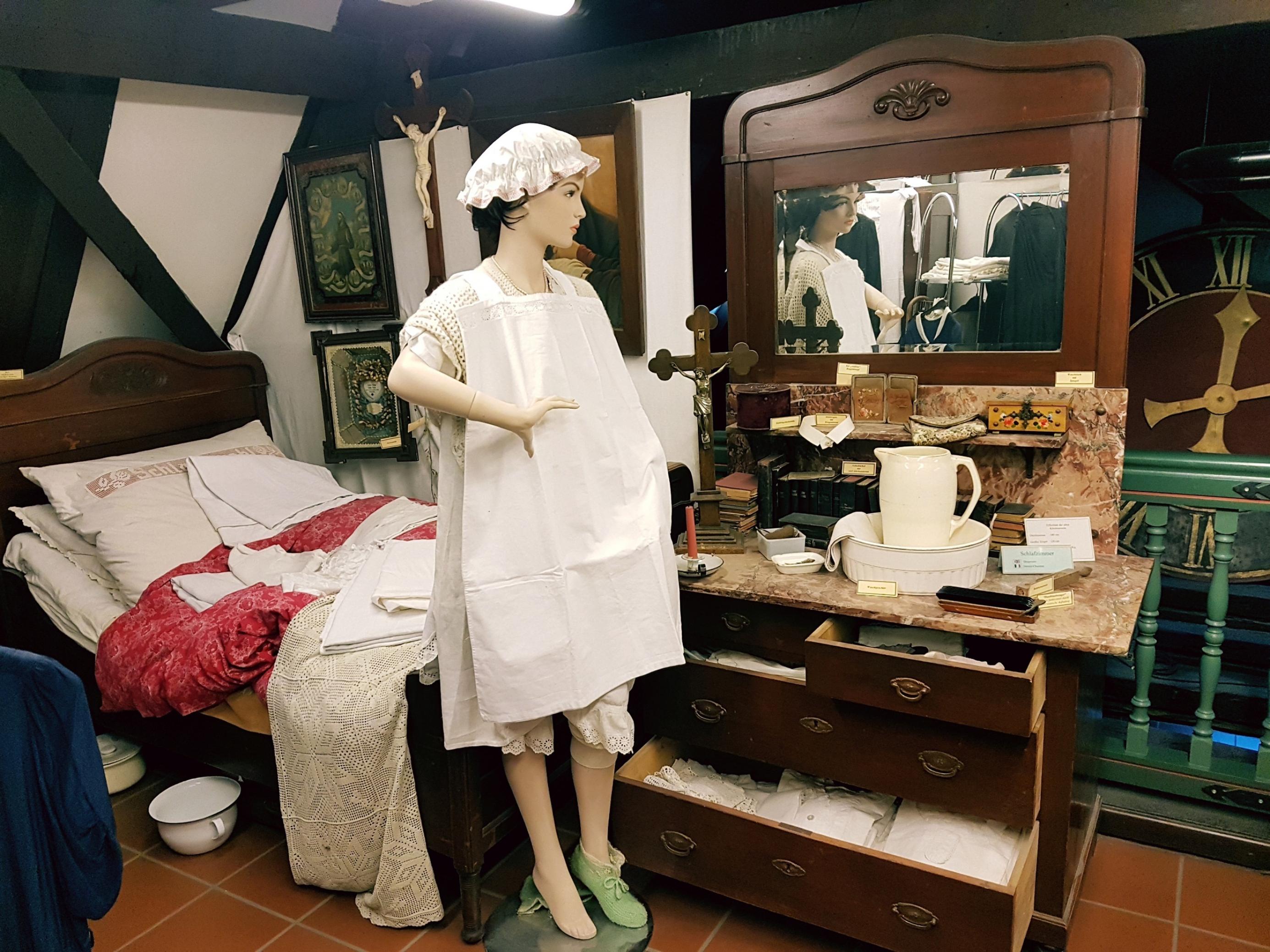 Eine Puppe mit alten Kleidern vor einem alten Spiegel, im Hintergrund ein Bett mit weißer Wäsche.