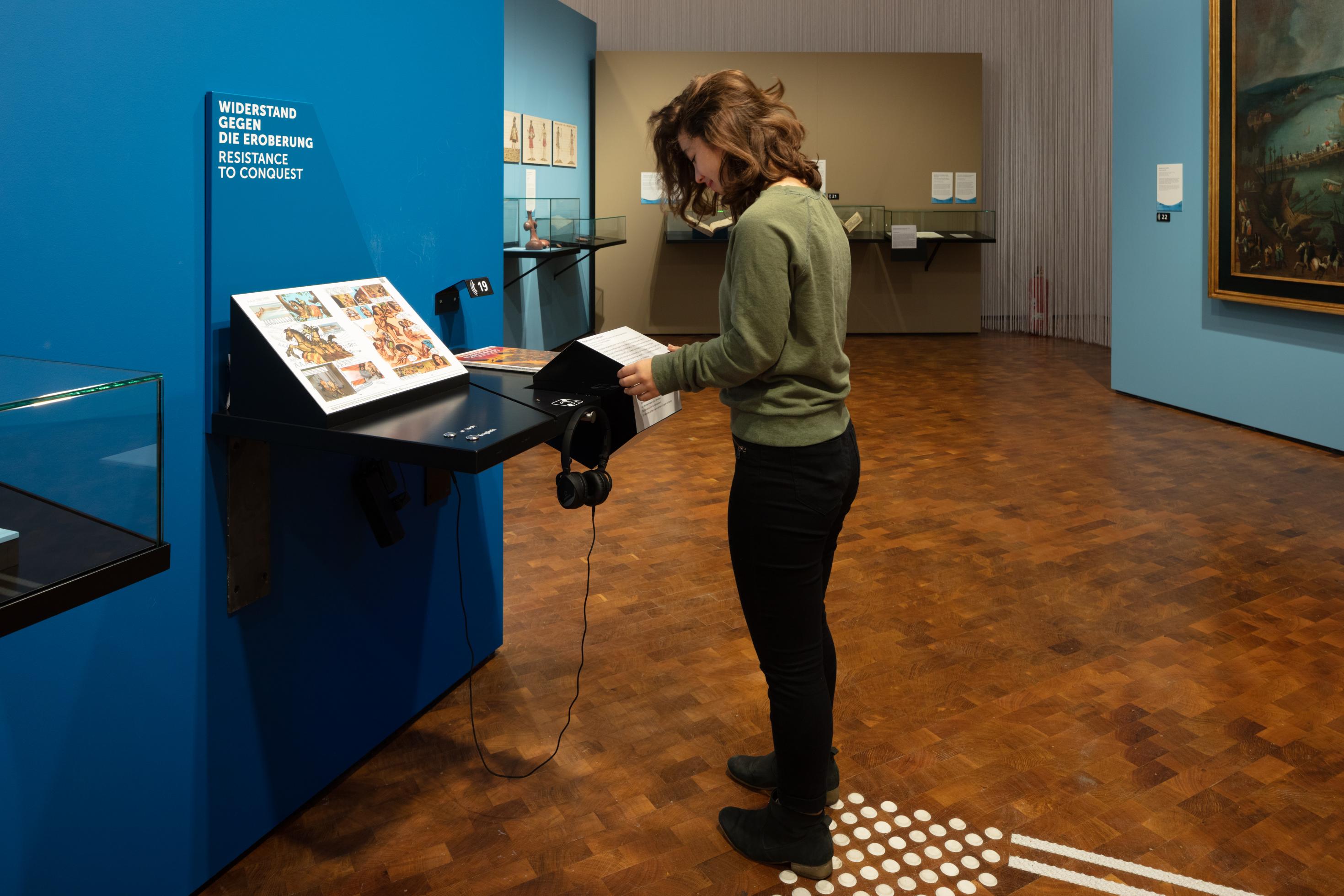 Eine junge Frau steht im Museum vor einer Info-Station und dreht an einer Trommel, die Informationen in verschiedenen Formen anbietet.