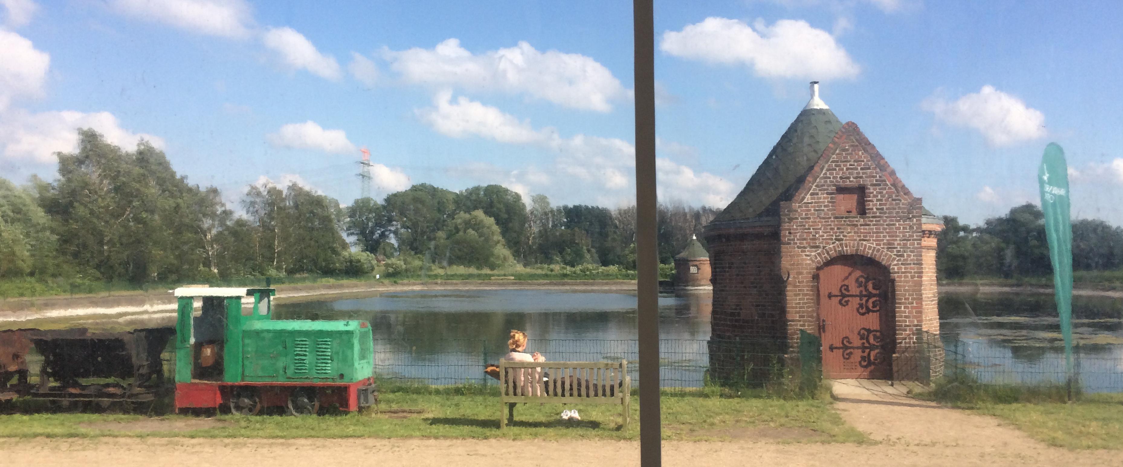 Blick über ein Wasserreservoir, davor ein Backsteinhäuschen und eine Grubenlokomotive. Ein Mensch sitzt auf einer Bank.