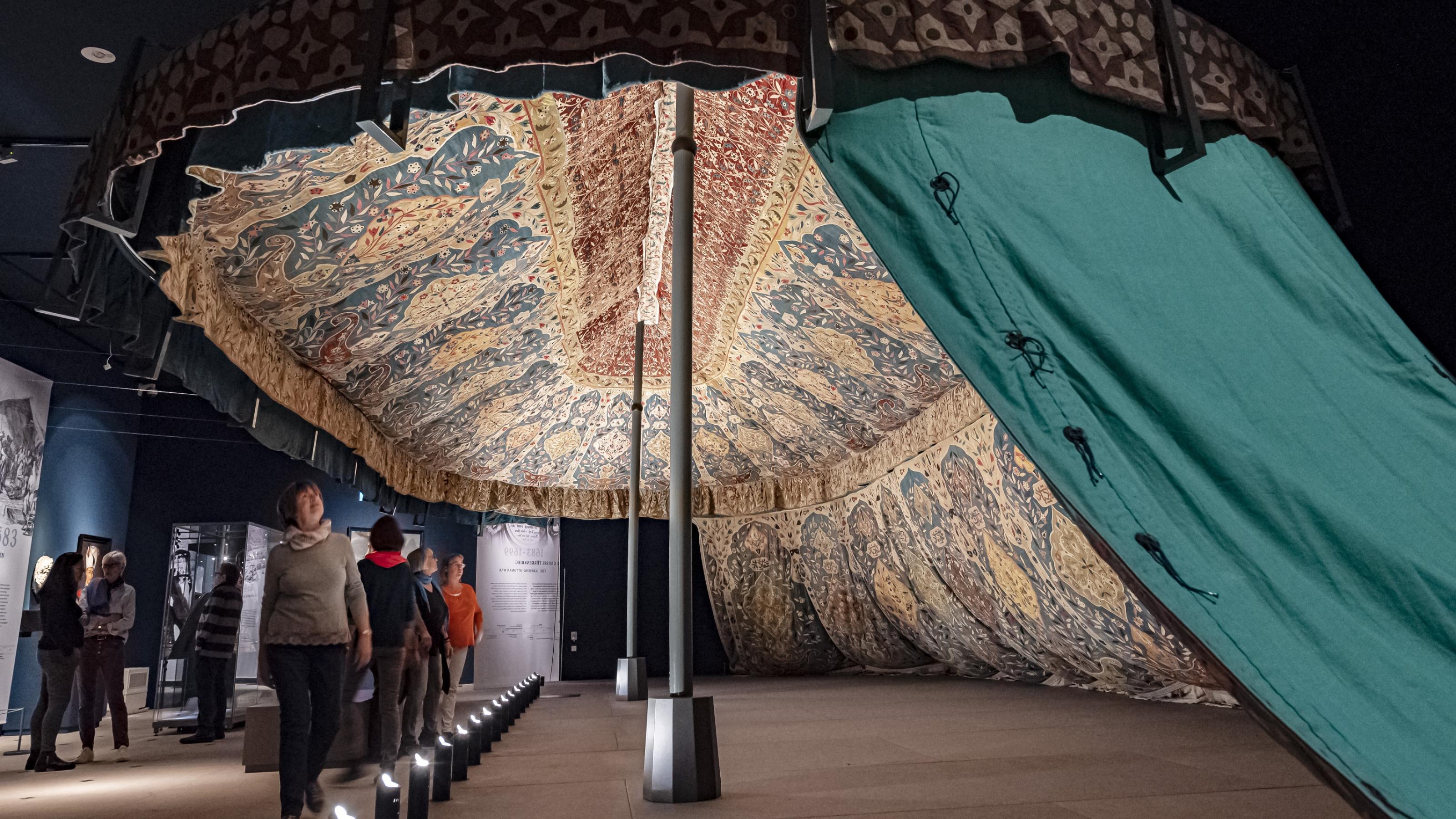 Ein zwei-mastiges, mit Ornamenten geschmücktes Zelt, deren eine Längsseite geöffnet ist.