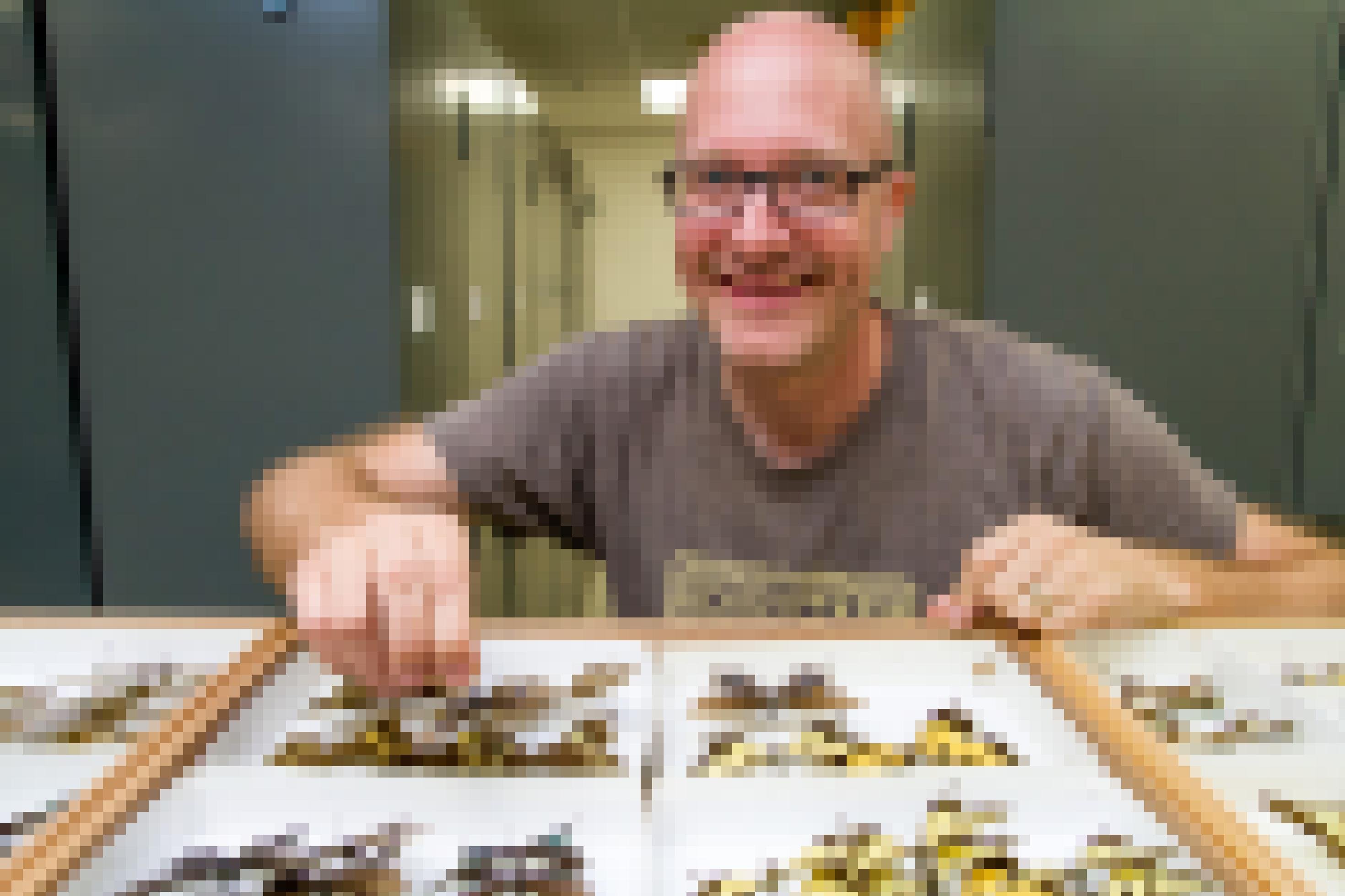 Ein kahler Mann mit Brille lächelt in die Kamera. Vor ihn liegen Kästen mit großen bunten Schmetterlingen.