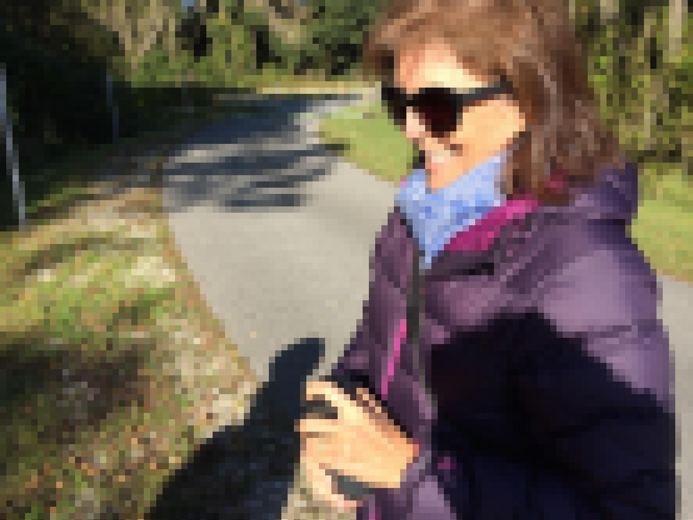 Eine Frau mit Sonnenbrille, schulterlangem braunen Haar und einer Daunenjacke steht lächelnd in der Sonne. In der Hand hält sie einen kleinen schwarzen Lautsprecher.