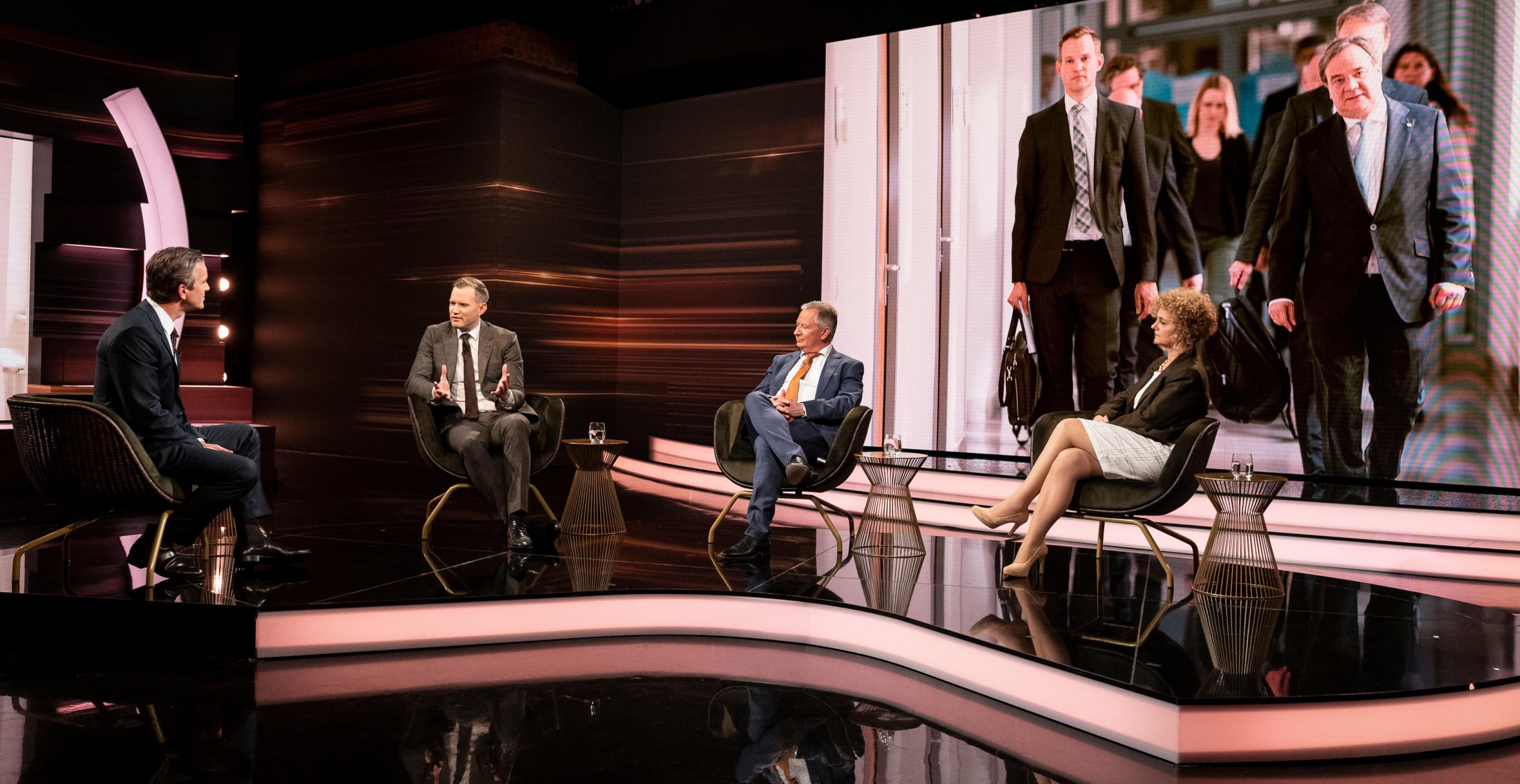 Drei Männer und eine Frau sitzen in einem in Brauntönen gehaltenen Fernsehstudio und sprechen miteinander. Im Hintergrund ein großes Foto, auf dem Menschen in Anzägen in Zweierreihen einen Korridor entlanggehen.