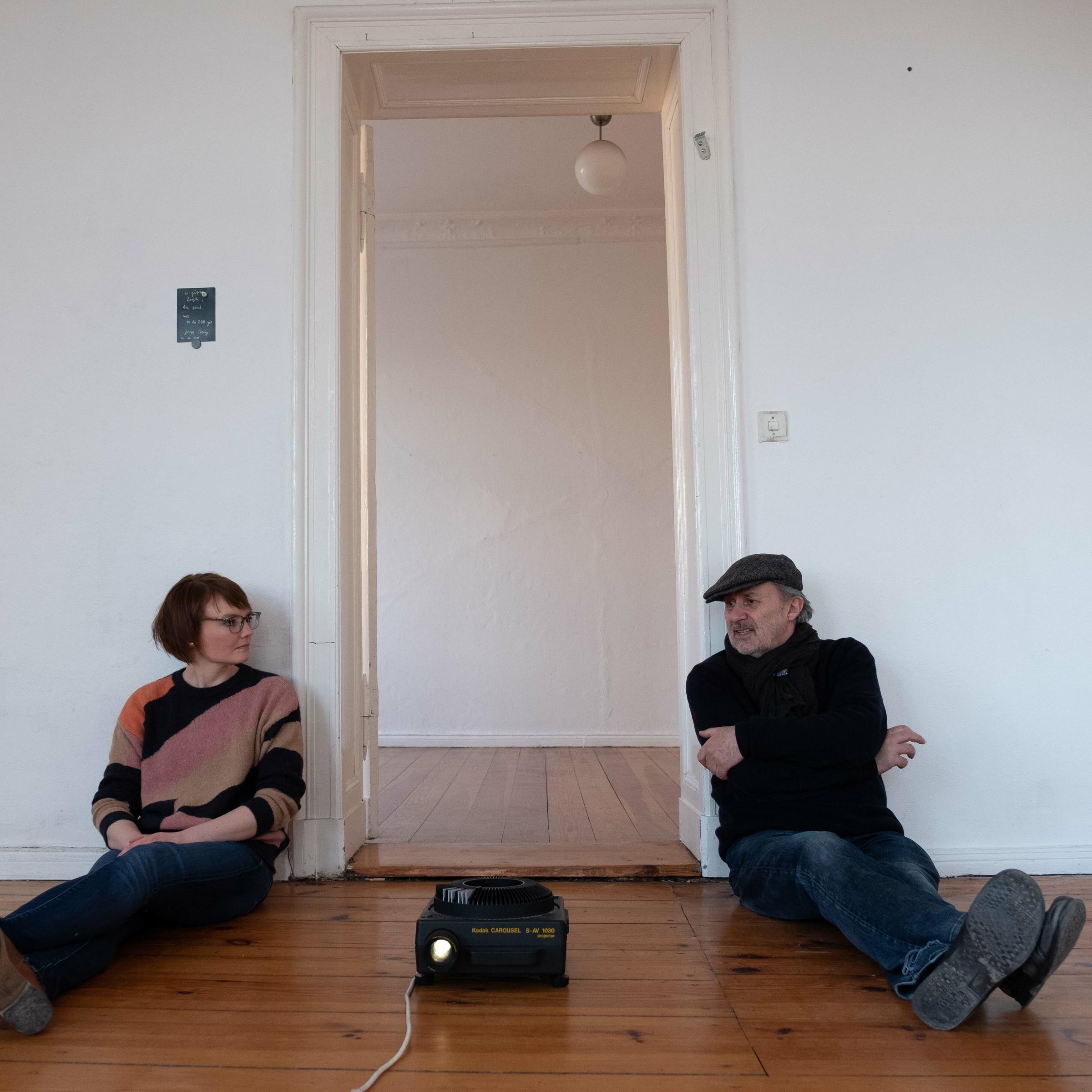 Sarah Alberti interviewt einen älteren Mann. Sein Name ist Grischa Meyer. Beide sitzen auf dem Fußboden einer leeren Wohnung.