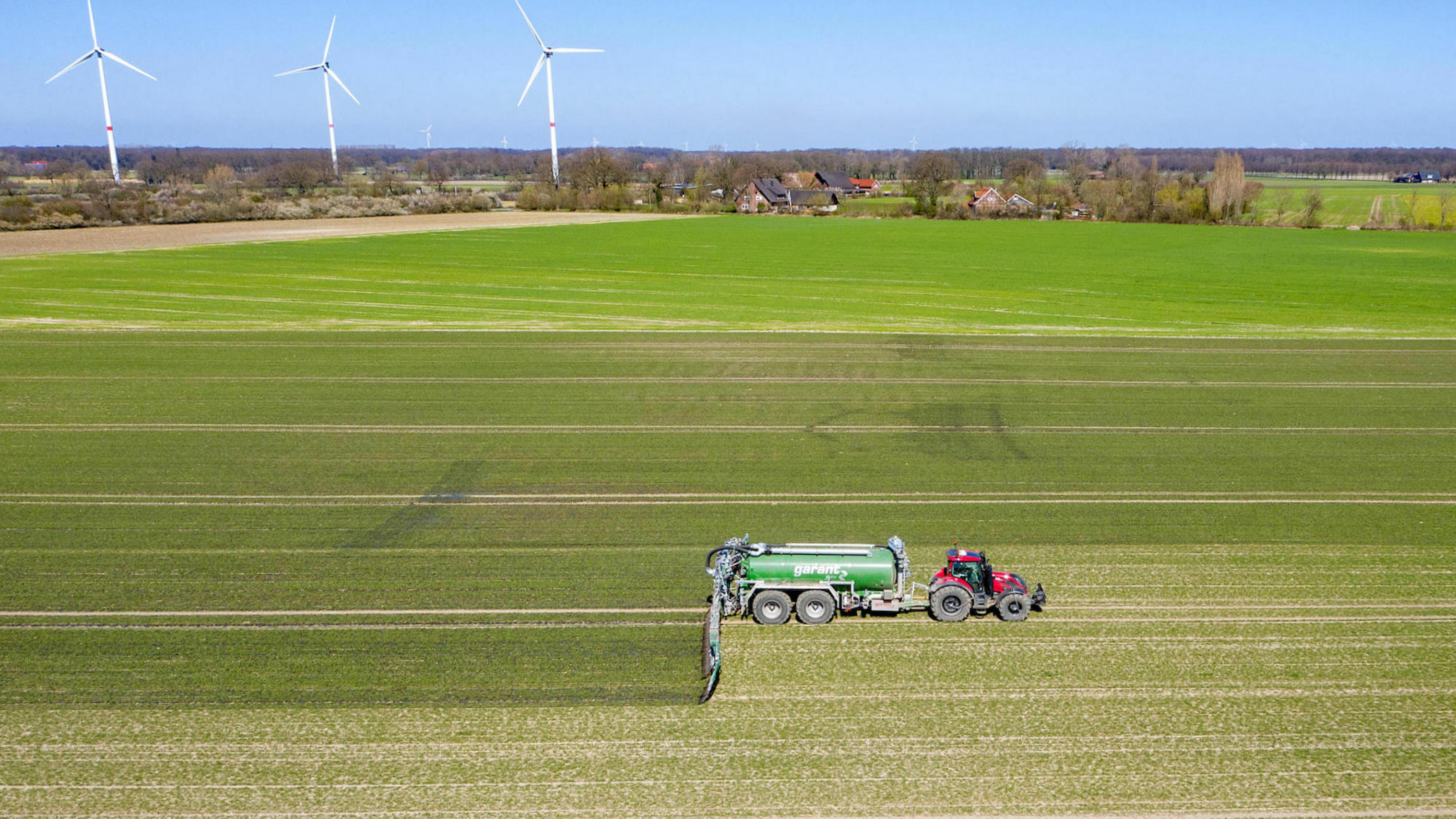 Luftaufnahme: Ein Trecker mit Güllefass bei der Ausbringung von Flüssigmist zur Düngung eines jungen Getreidebestandes im Frühjahr, im Hintergrund drei Windräder.