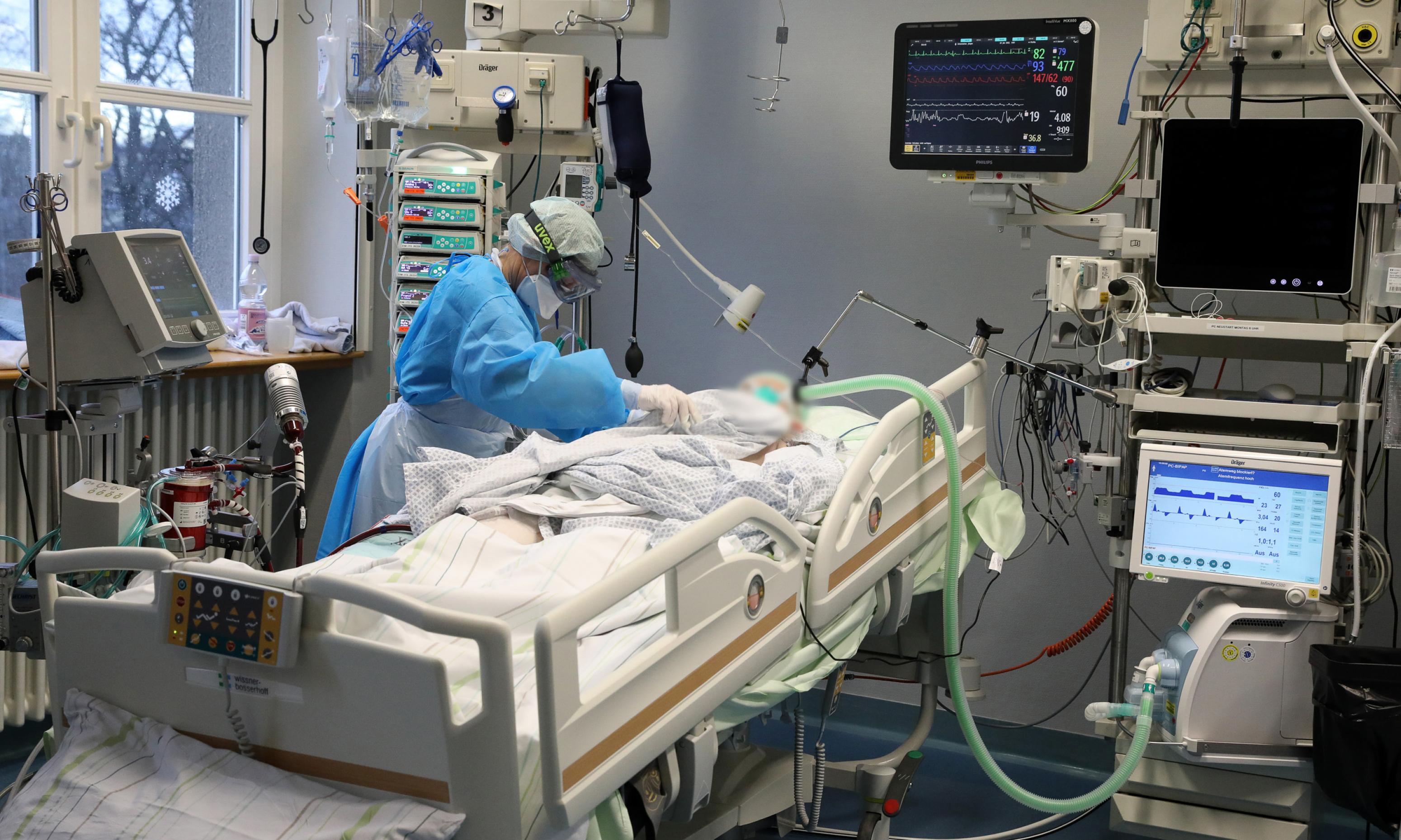 Ein Krankenbett mit einem Patienten in weißem Hemdchen. Rundherum medizinische Geräte mit Bildschirmen. Eine Person in blauem Schutzanzug mit Schütze, Gesichtsmaske, Haube, Handschuhen und Skibrille beugt sich über den Patienten.