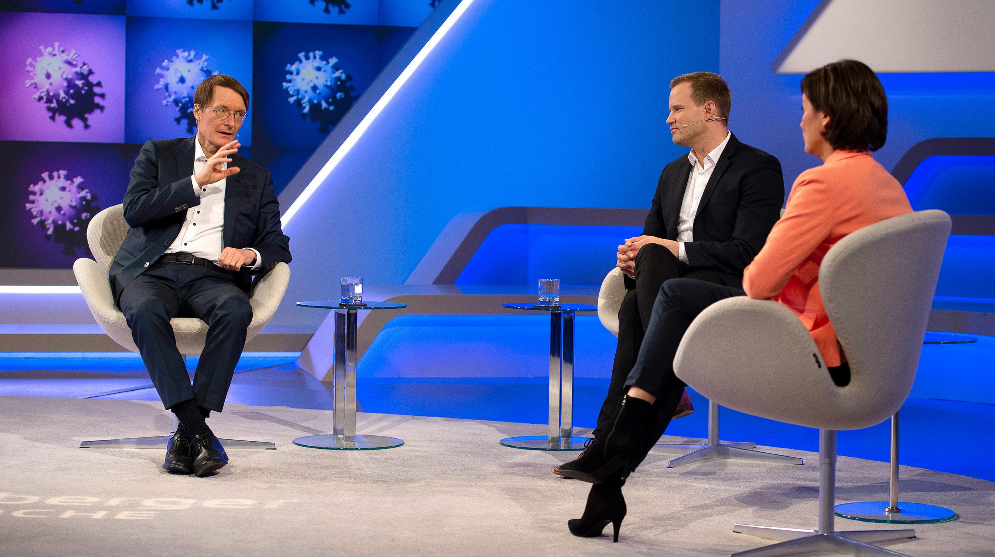 Zwei Männer in Anzügen und eine Frau im lachsfarbenen Blazer sitzen in einem in Blautönen gehaltenen Fernsehstudio. Im Hintergrund sind Bilder des Sars-Coronavirus-2 an der Wand.