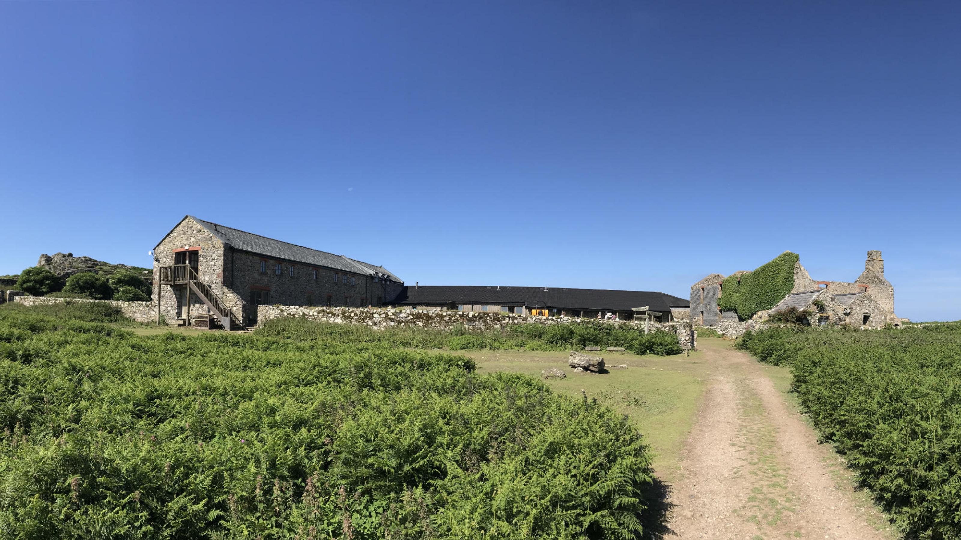 Ein unbefestigter Weg führt auf einen Hof zu, rechts eine alte Ruine, links ein zweistöckiges Gebäude aus Naturstein. Rechte: Anne Preger