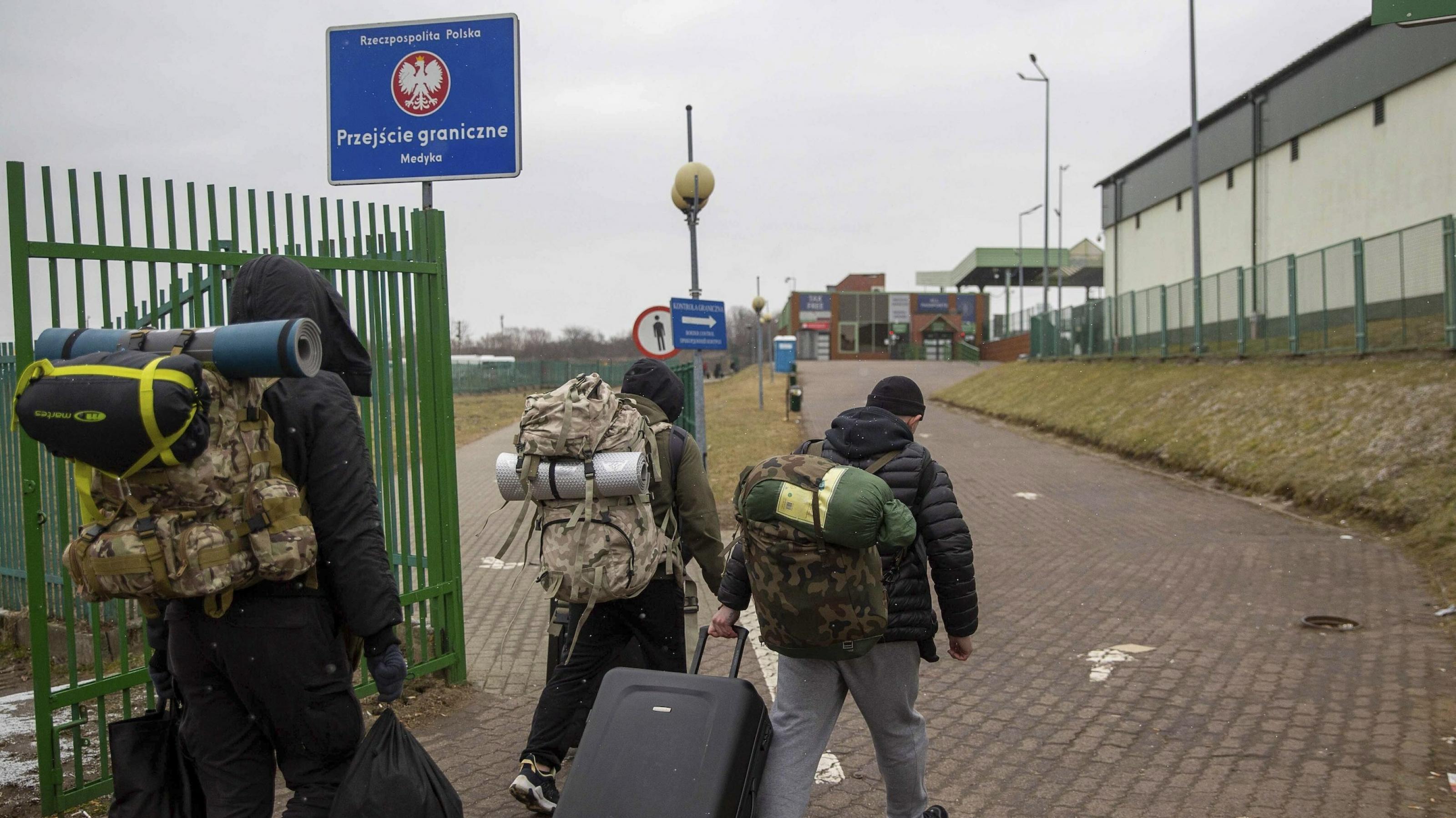 Drei Männer mit Rucksäcken und Koffern von hinten gesehen in einer Grenzanlage, die man an einem Schild mit dem polnischen Hoheitszeichen erkennt.