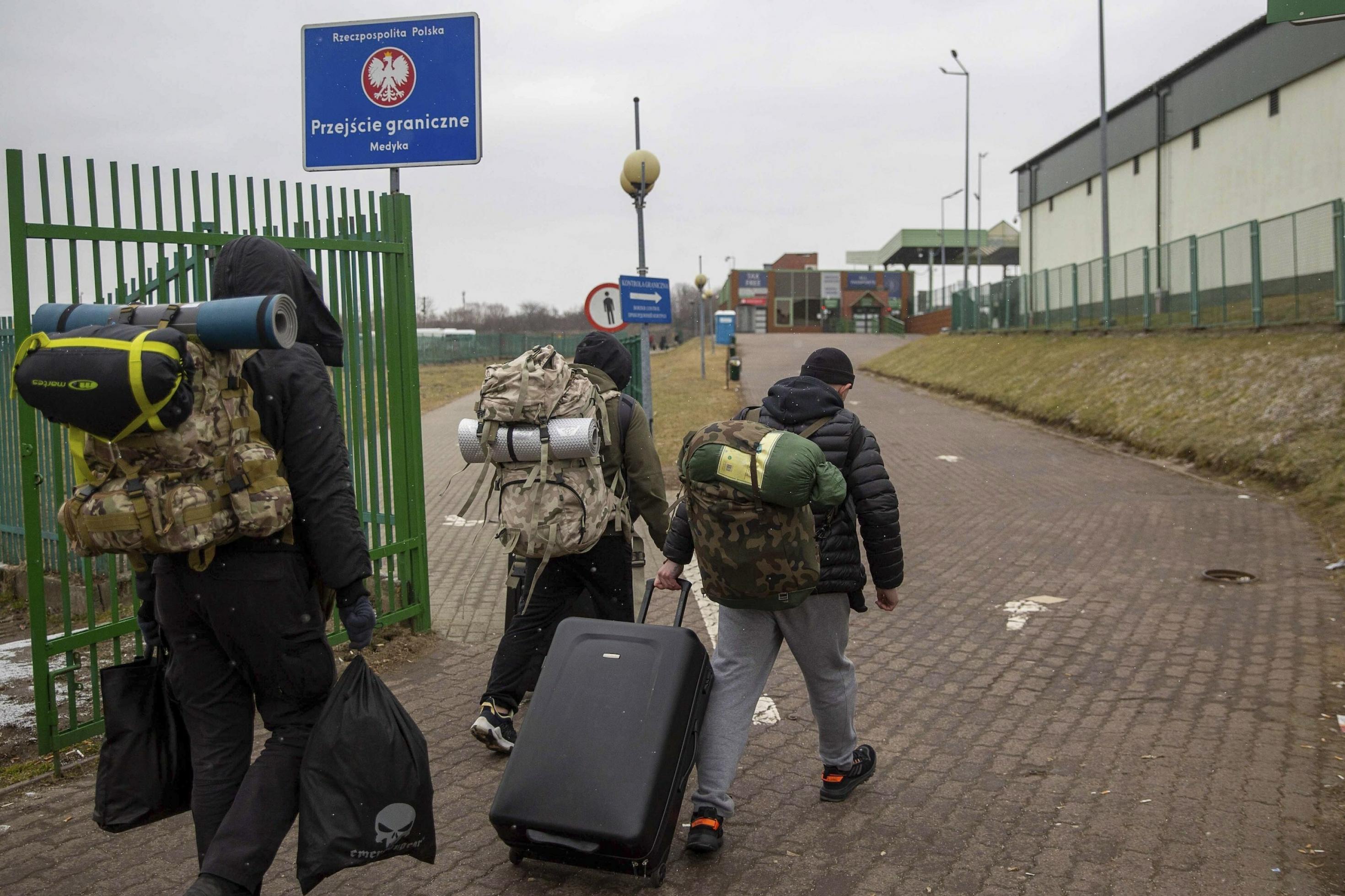 Drei Männer mit Rucksäcken und Koffern von hinten gesehen in einer Grenzanlage, die man an einem Schild mit dem polnischen Hoheitszeichen erkennt.