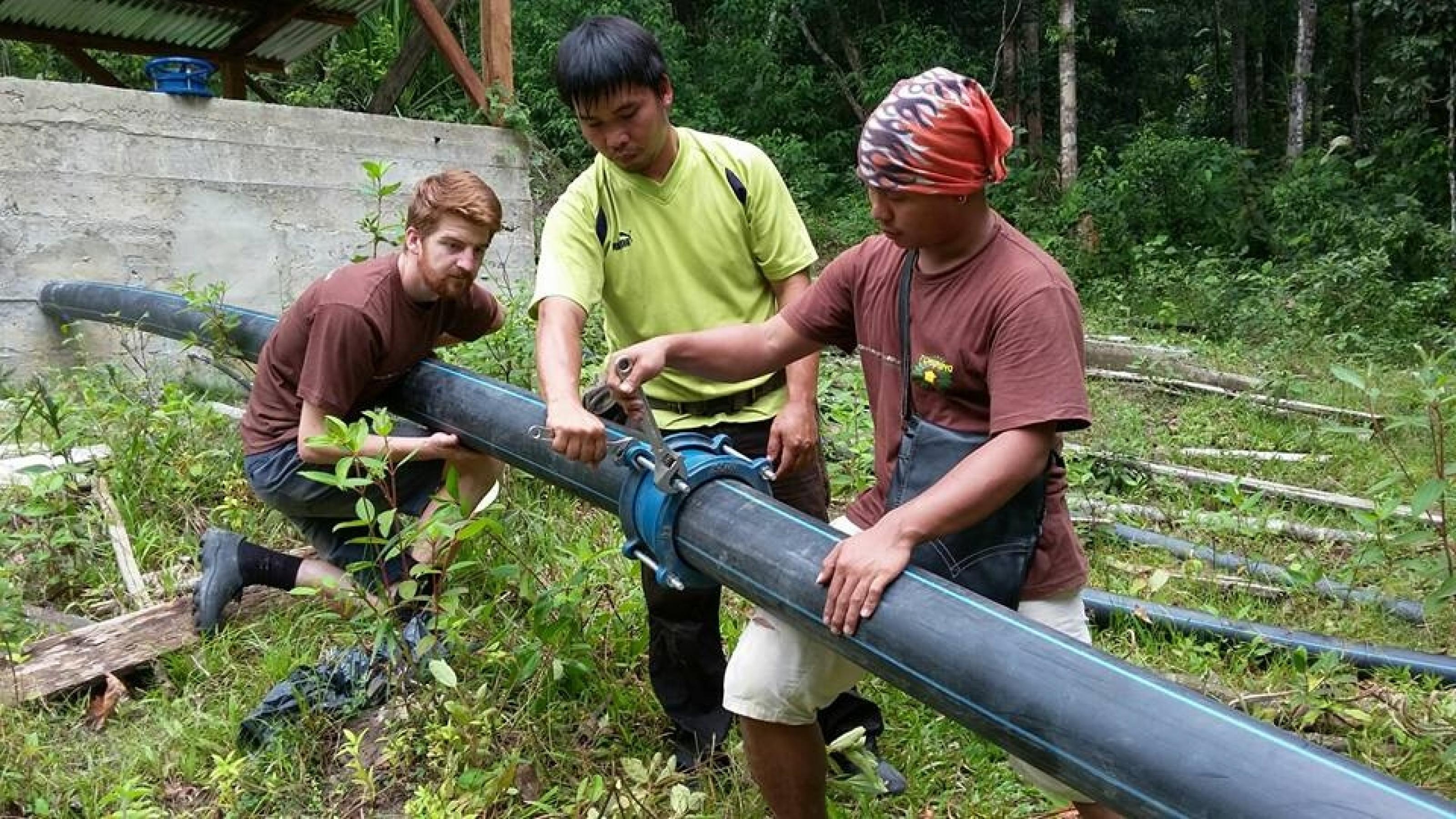 Das Foto zeigt drei Männer, die ein dickes schwarzes Wasserrohr verlegen. – 
… Am Ort werden dann dicke Plastikrohre verlegt, durch die das Wasser zur Turbine strömt.Oft sind freiwillige Helfer aus westlichen Staaten dabei. …