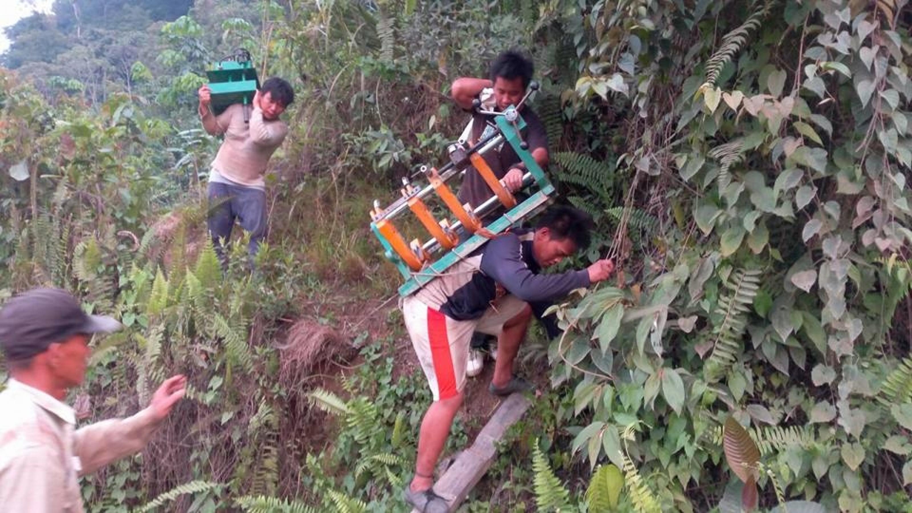 Das Foto zeigt mehrere Männer, die Maschinenteile auf dem Rücken über einen einfachen Pfad tragen und einen Anstieg mittels einer Leiter überwinden. – 
… Oft genug müssen die Teile des neuen Wasserkraftwerks auf dem Rücken transportiert werden. …