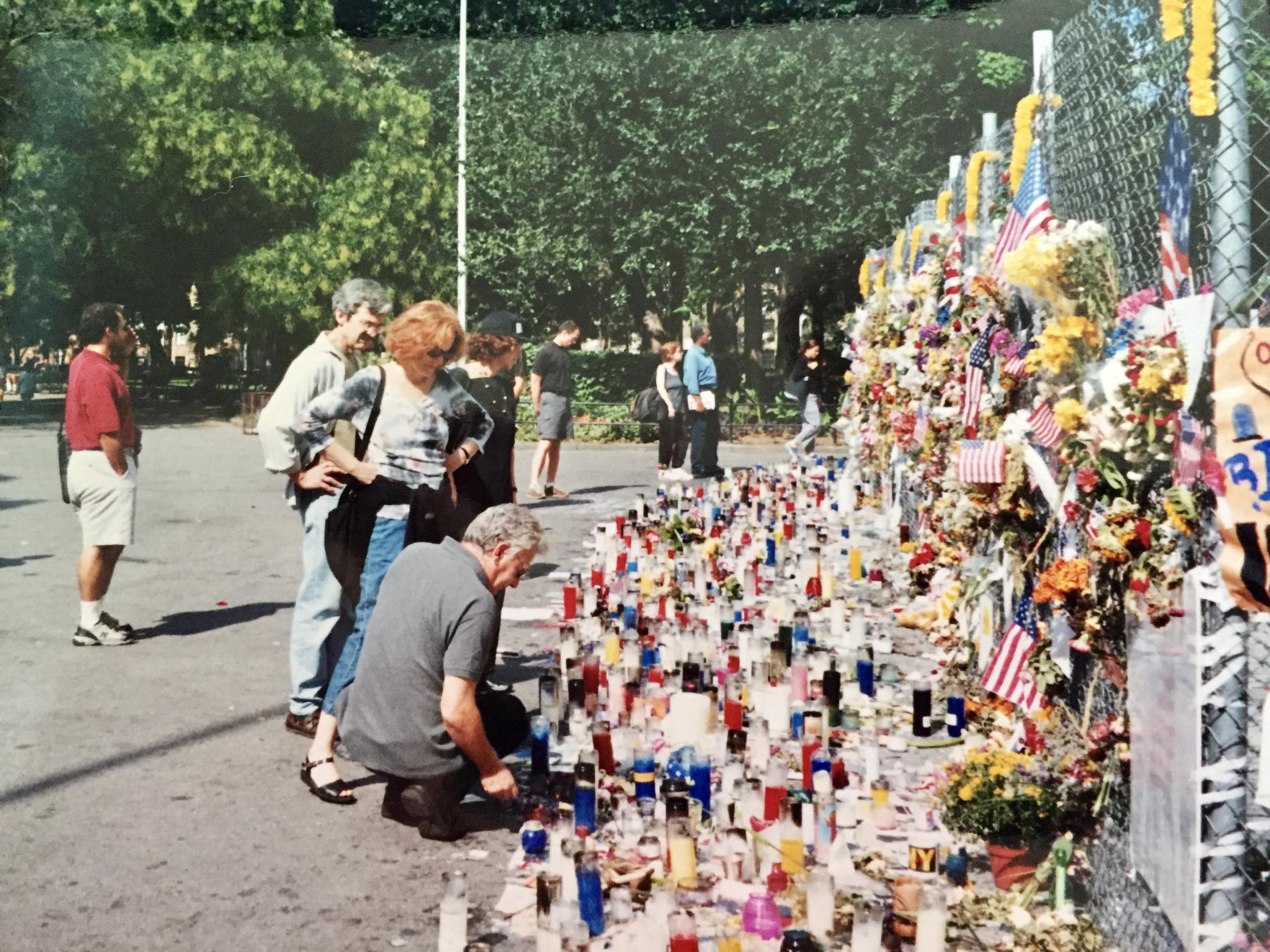 Männer und Frauen stehen vor einem Maschendrahtzaun, wo Kerzen, Fahnen und Kränze an die Opfer der Anschläge vom 11. September erinnern. Im Hintergrund Bäume und ein Park
