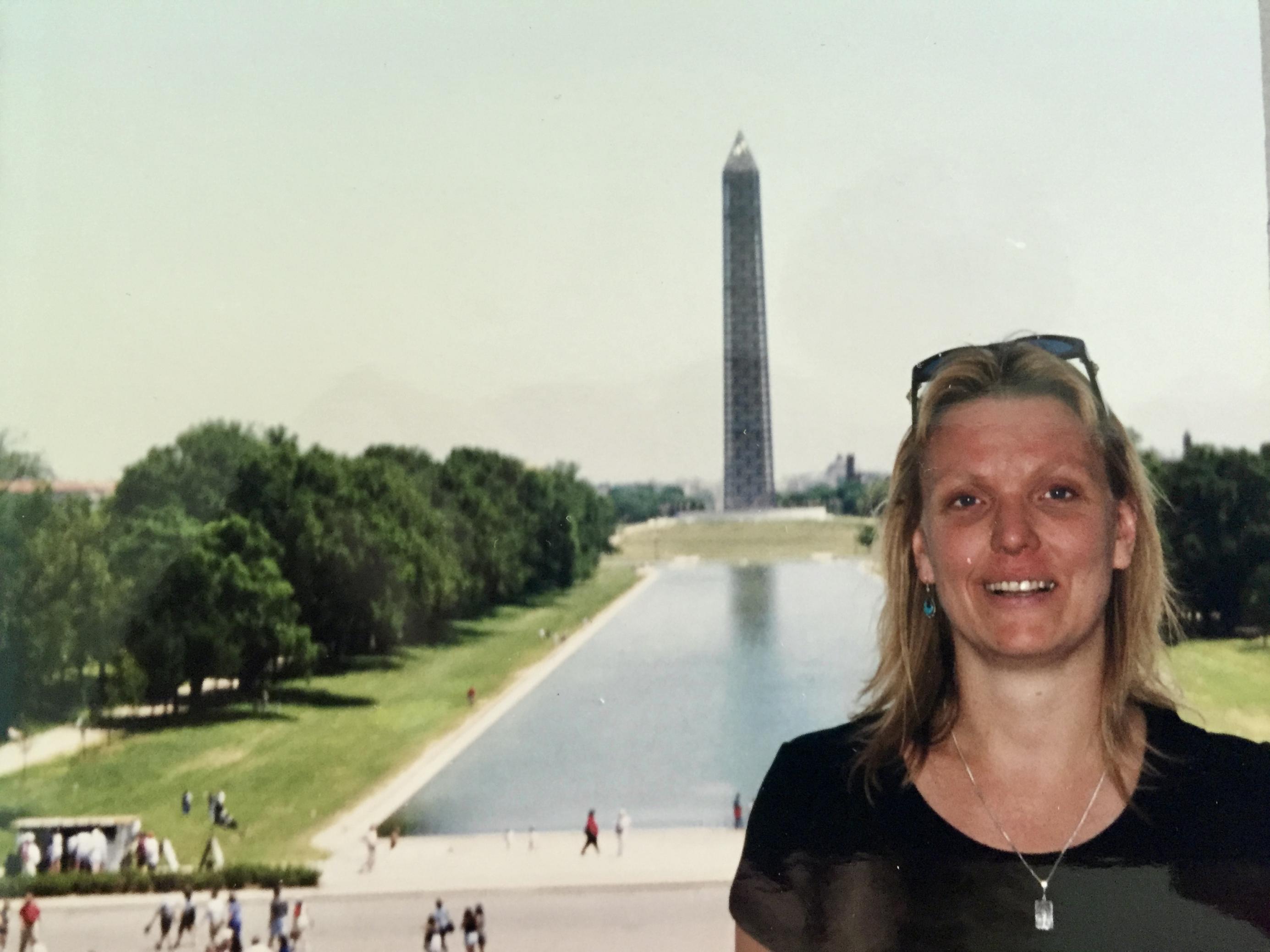 Eine Frau Mitte dreißig, blond mit dunklem T-Shirt, posiert vor dem Reflecting pool und dem Washington Monument