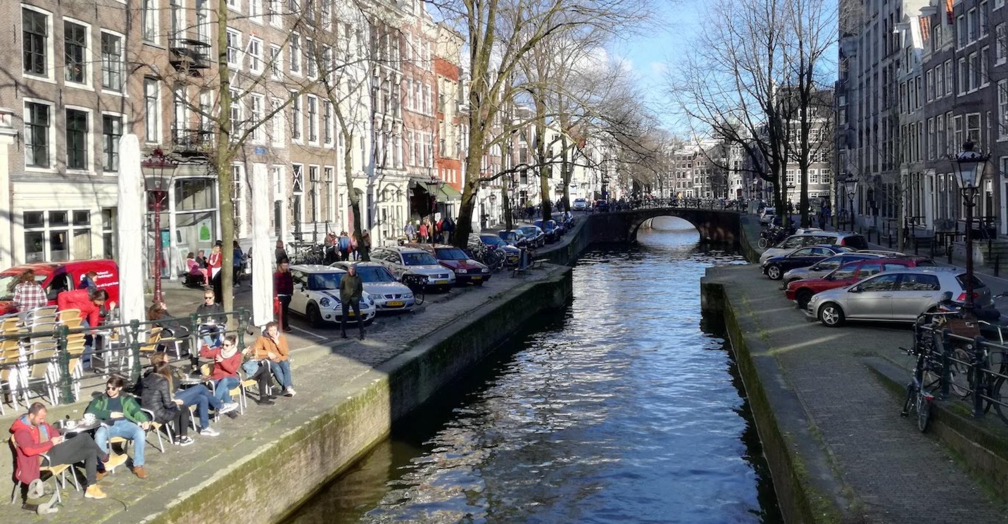 Blick von einer Brücke auf eine Gracht in Amsterdam, an der beidseitig Autos parken.