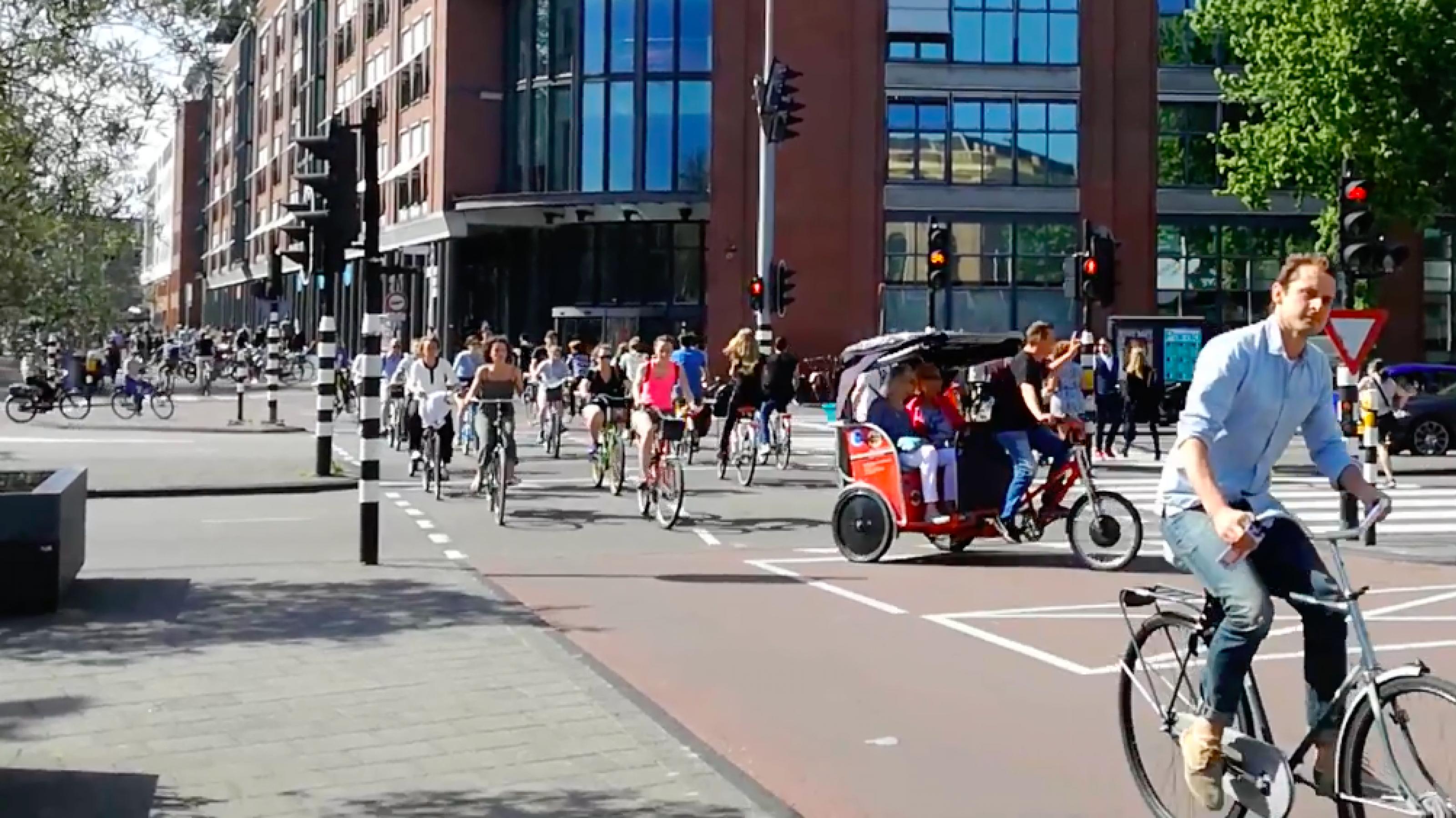 Ein Pulk Radfahrern und eine Rikscha queren eine Kreuzung in Amsterdam