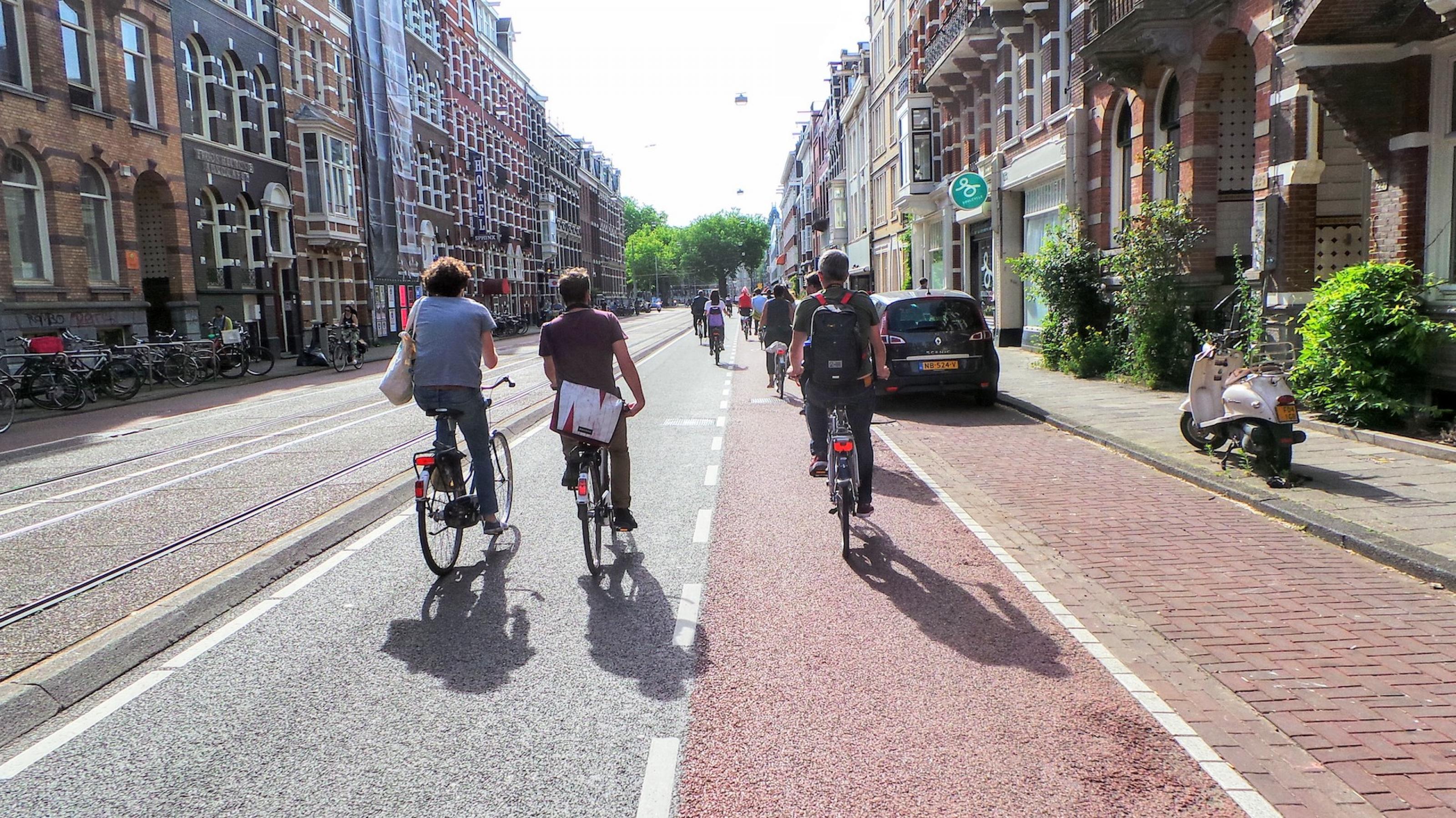 Von hinten sind drei Radfahrer im Vordergrund zu sehen, wie sie mitten auf einer Straße fahren.