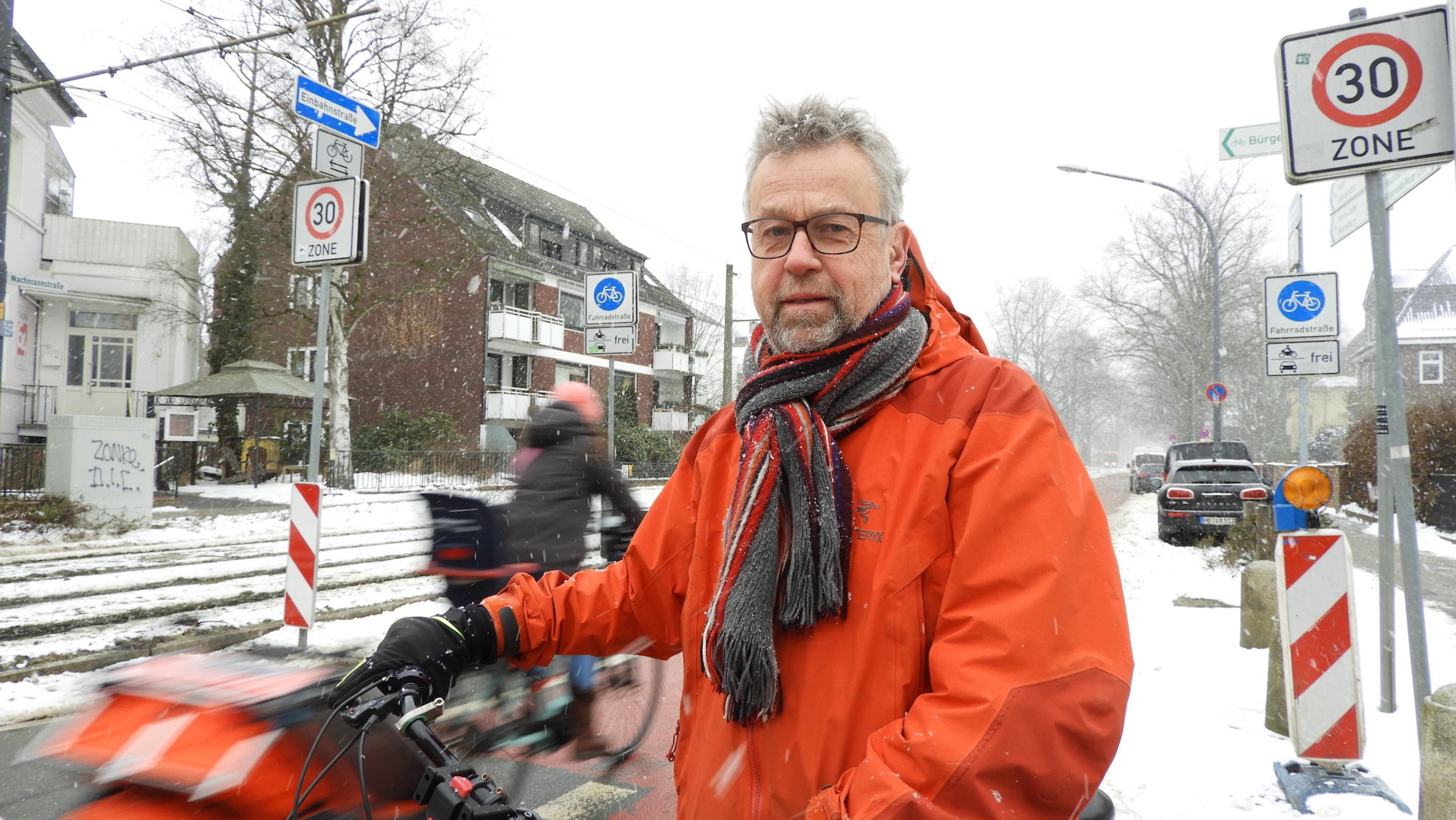 Michael Glotz-Richter steht vor einer Fahrradstraße. Er trägt eine rote Jacke und hat graue lockige Haare, einen Bart und trägt eine Brille