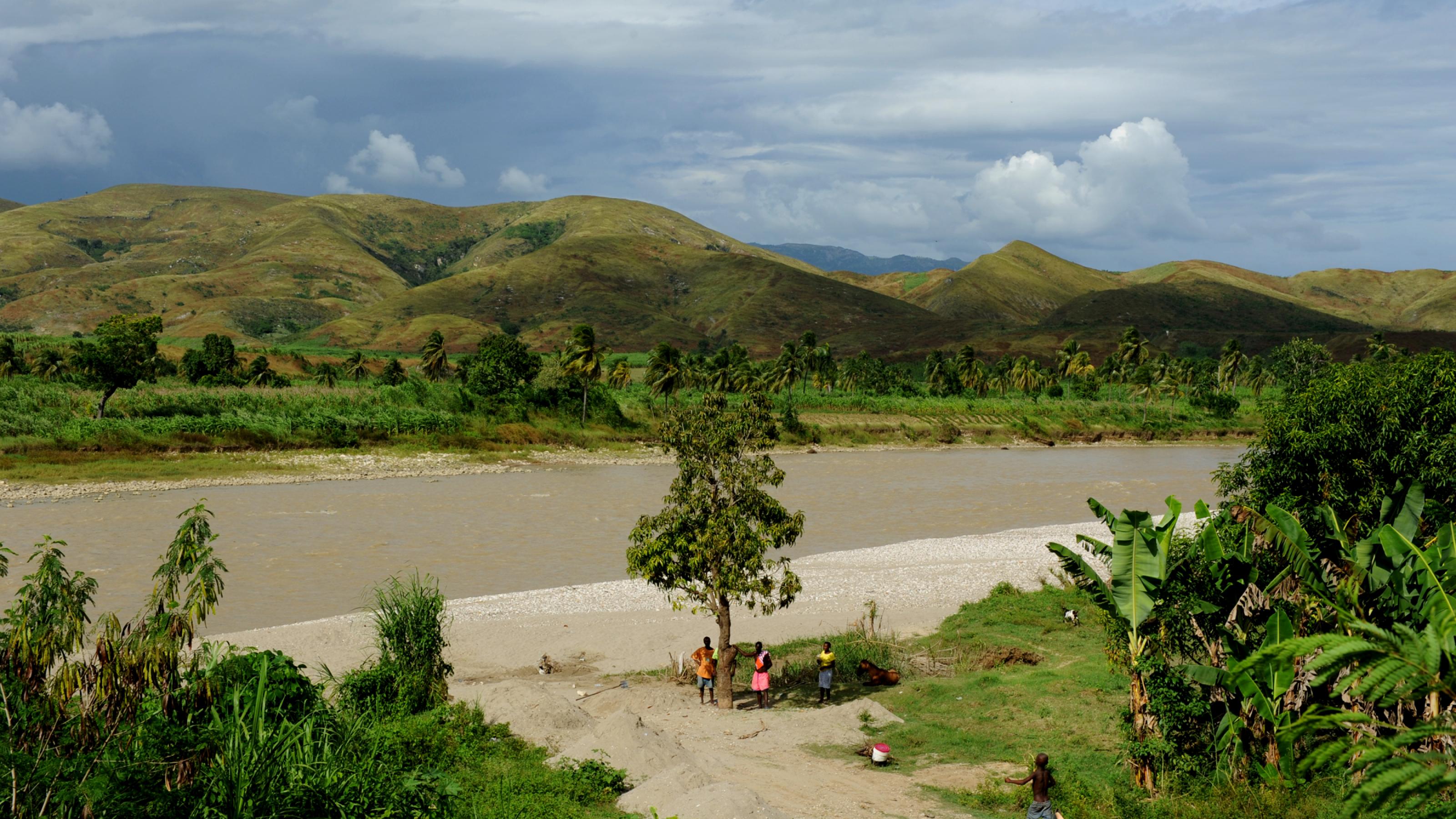 Das Bild zeigt eine Flusslandschaft auf Haiti. Vor kahlen, grünen Bergen fließt ein mittelgroßer Fluss, der Artibonite. Einige Menschen sind auch im Bild, es ist nicht klar, was sie machen.