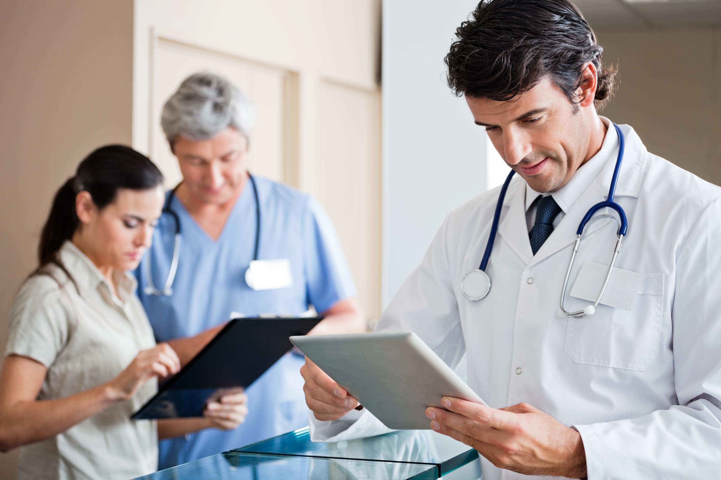 Ein Arzt steht in einer Praxis und schaut auf ein Tablet, im Hintergrund sind zwei weitere Gesundheitsfachkräfte zu sehen.