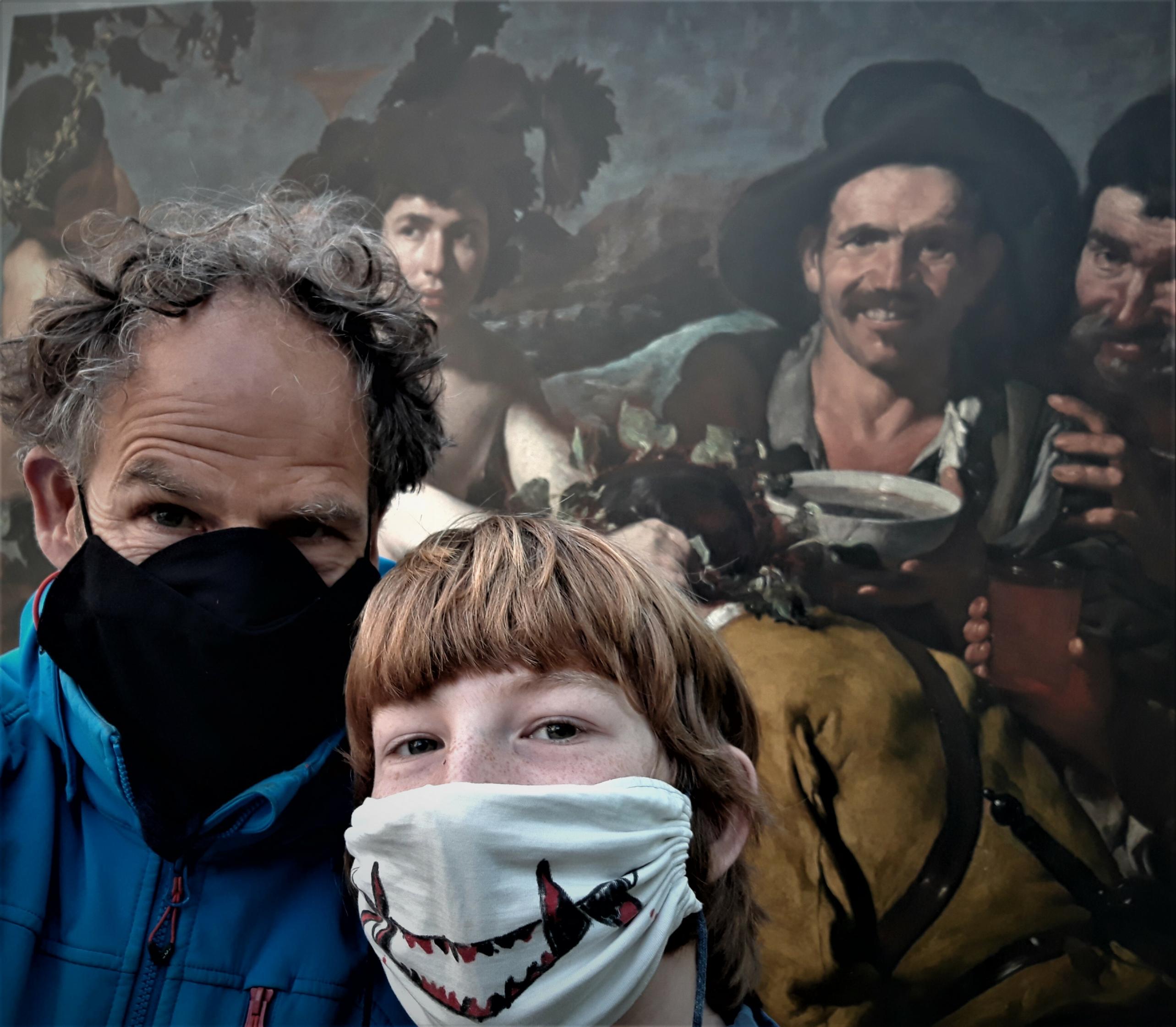Vater und Sohn – beide mit Anti-Corona-Masken – posieren vor einem Ausschnitt des Gemäldes, auf dem in erster Linie der eine weiße Schale haltende „Trinker“ zu sehen ist und rechterhand von diesem der Kopf Bacchus'.