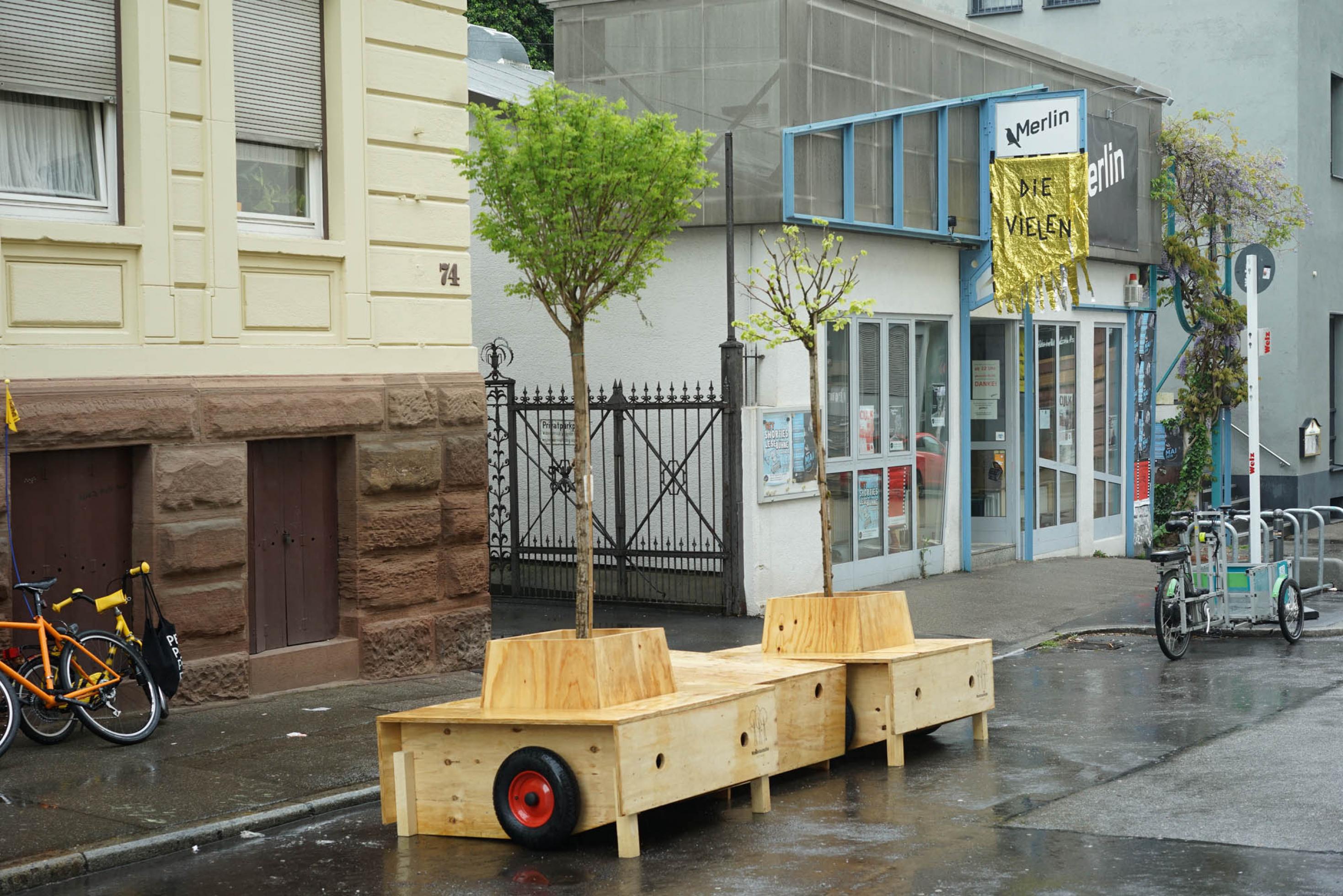 Junge Bäume mit einer Holzeinfassung, die eine Sitzgelegenheit darstellt, stehen am Straßenrand.