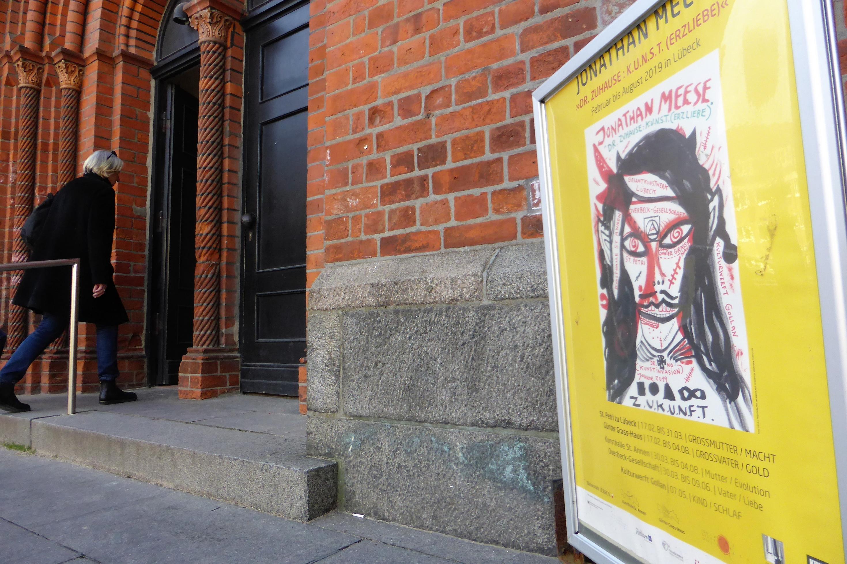 gelbes Plakat, rechts vor dem Eingangsportal der Backsteinkirche.