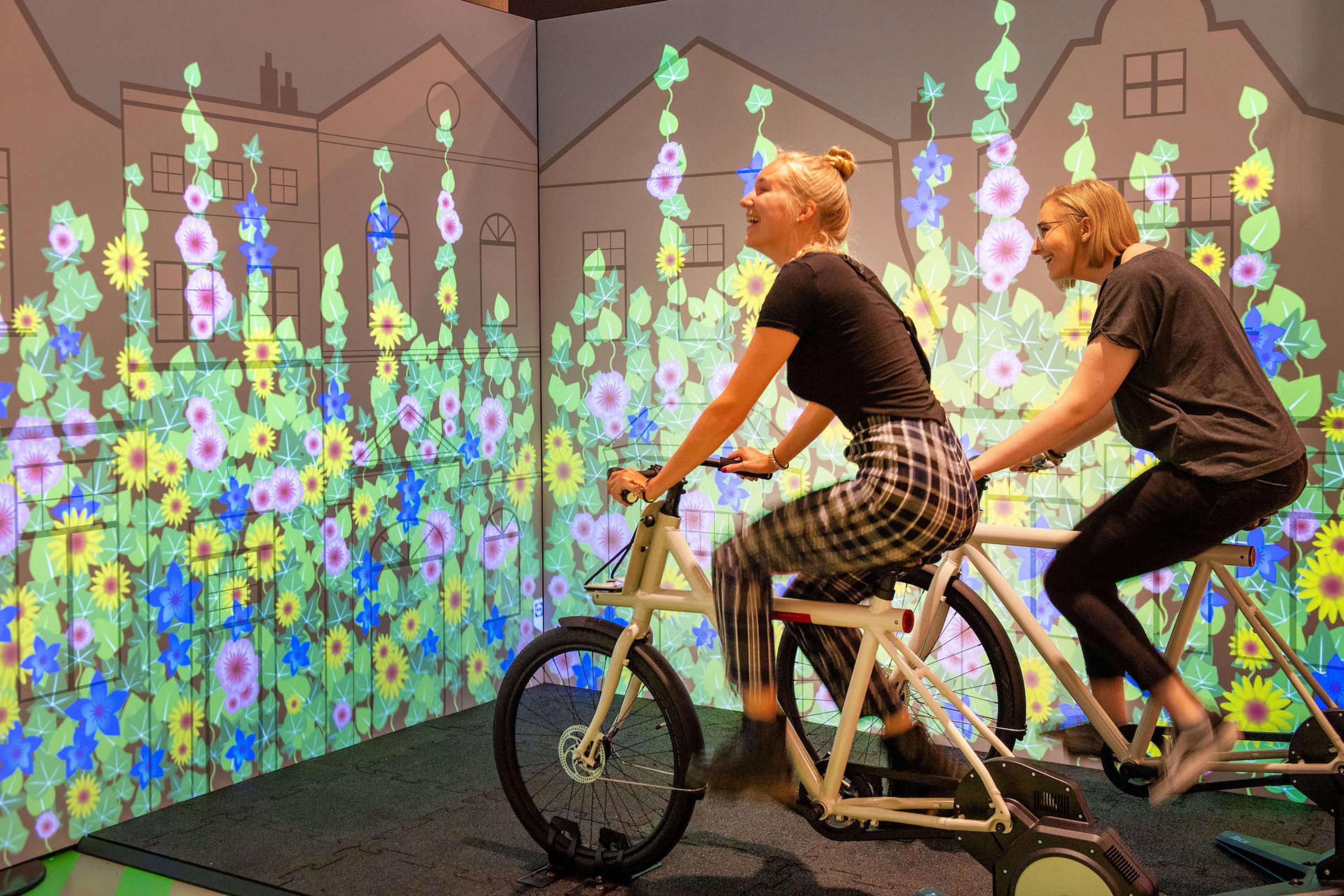 In einer Ecke der Ausstellung treten zwei Frauen auf weißen Fahrrädern schnell in die Pedale. Je schneller sie in die Pedale treten umso  mehrvirtuelle Blumen ranken sich die Wand empor
