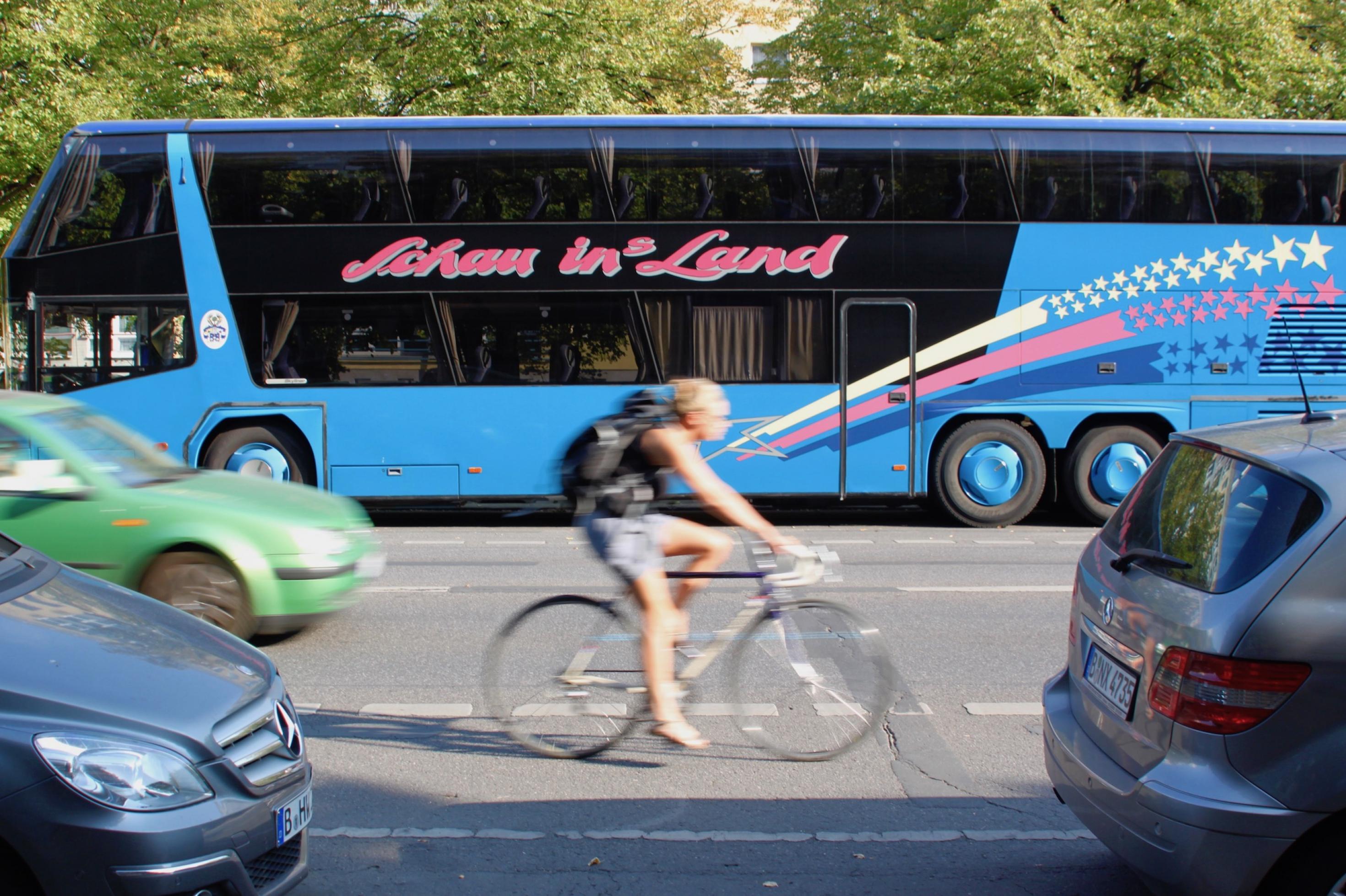 Es ist Sommer. Das Bild zeigt, die leicht verschwommen Seitenansicht einer Radfahrerin, die in T-Shirt und kurzer Hose auf einem blau-weißen Rennrad über die Straße fährt. Auf der gegenüberliegenden Seite parkt ein Bus mit der Aufschrift: Schau ins Land.