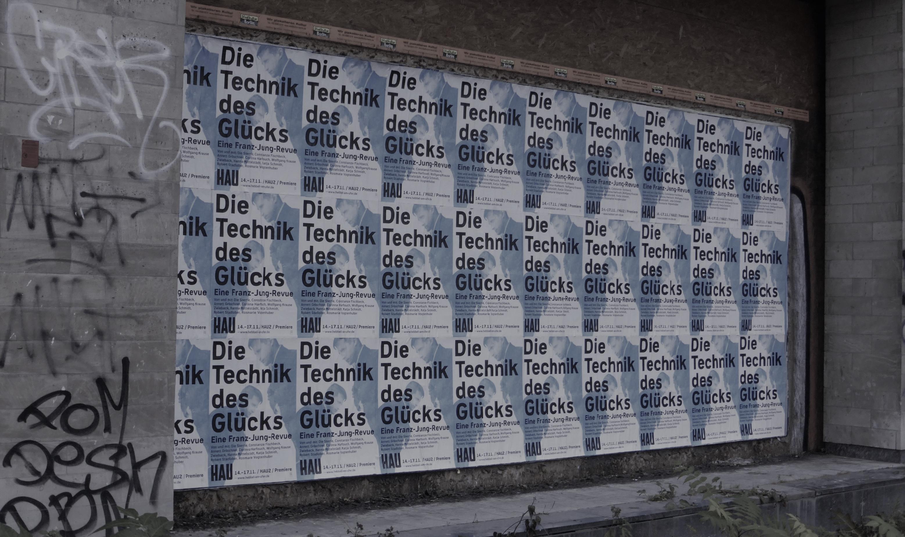 An der Bretterwand vor dem breiten, zugemauerten Eingang zu dem riesigen Gebäudekomplex sind 3 auf 9 DIN-A-2-große Plakate mit immer derselben Werbeaufschrift für eine „Revue“-Veranstaltung aufgeklebt. Die fetten Lettern verheißen „Die Technik des Glücks“.