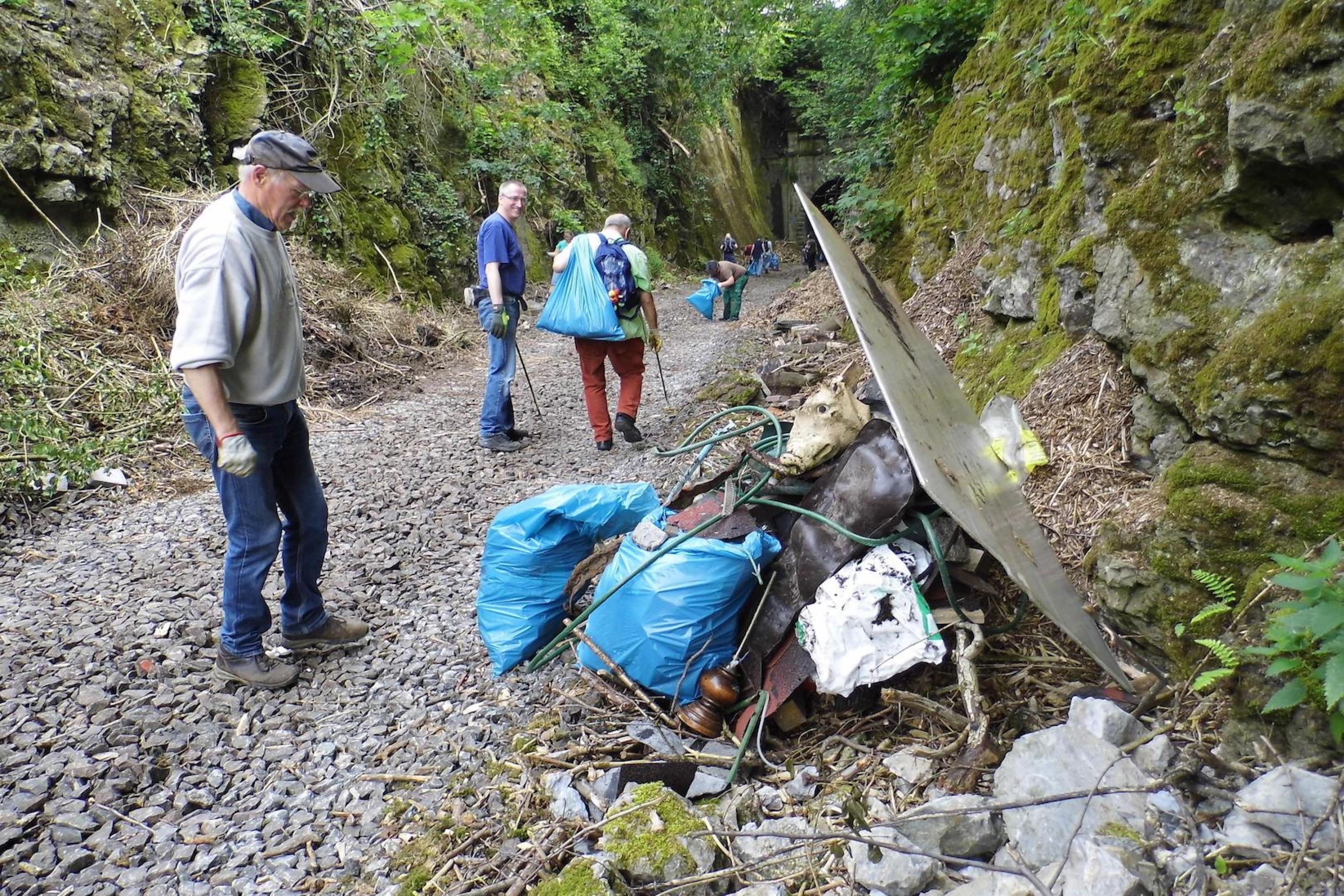 Vier ältere Menschen laufen entlang eines steinigen Weges und sammeln in blauen Säcken den Müll vom Wegesrand.