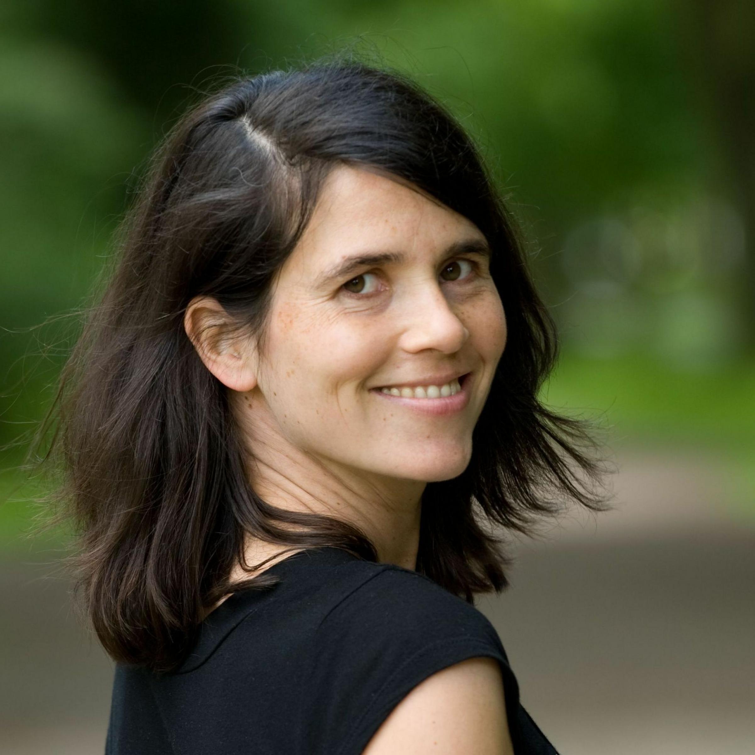 Das Foto zeigt die Journalistin und Buchautorin Carina Frey. Sie steht im  Park und schaut über die Schulter, das Grün der Bäume ist verschwommen im Hintergrund zu sehen.