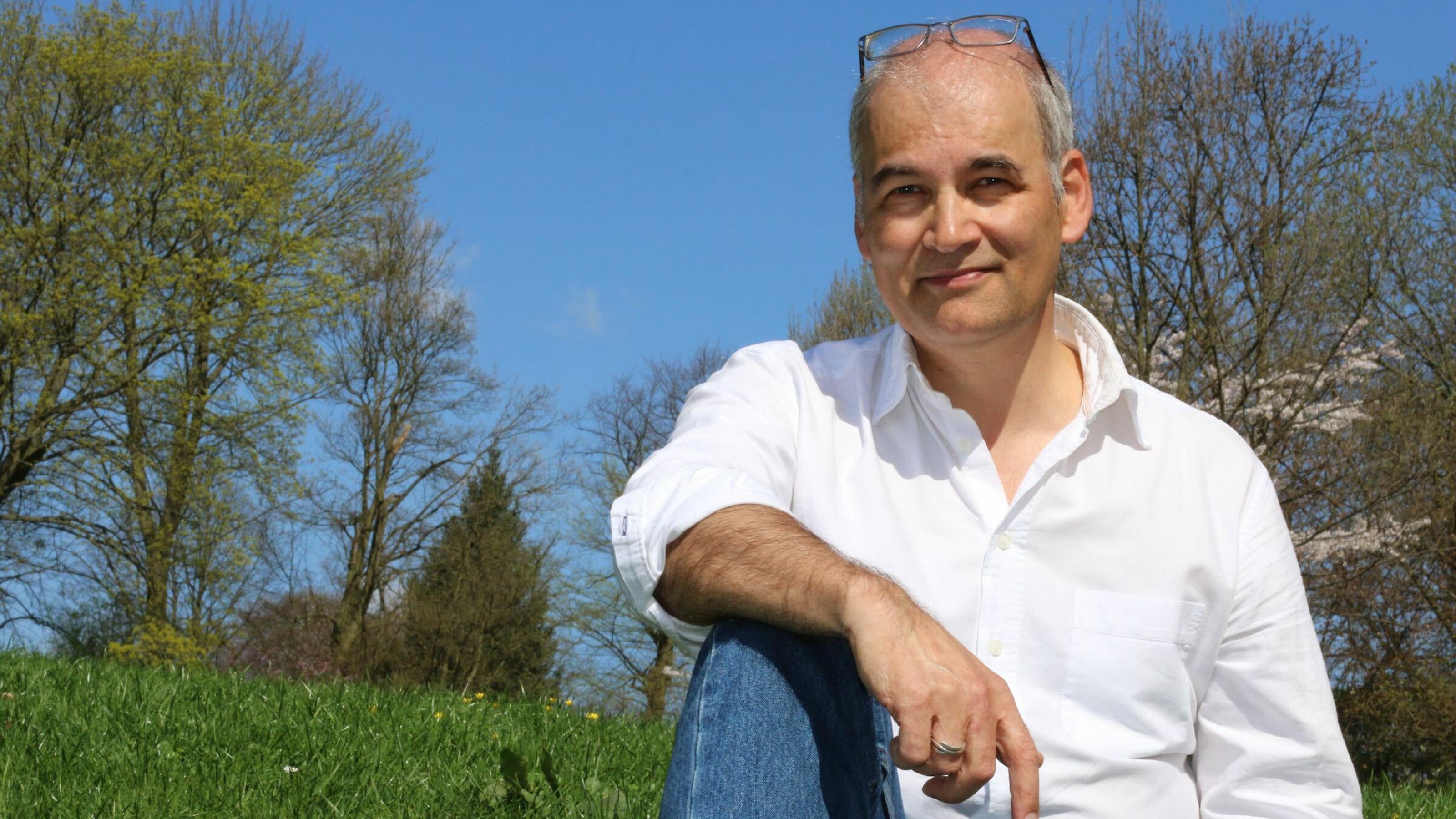 Der Journalist Christopher Schrader sitzt im weißen Hemd und Jeans auf einer Blumenwiese in einem Park am Elbhang. Er hat die Brille auf die Stirn geschoben, der rechte Arm liegt auf dem aufgestellten Knie.