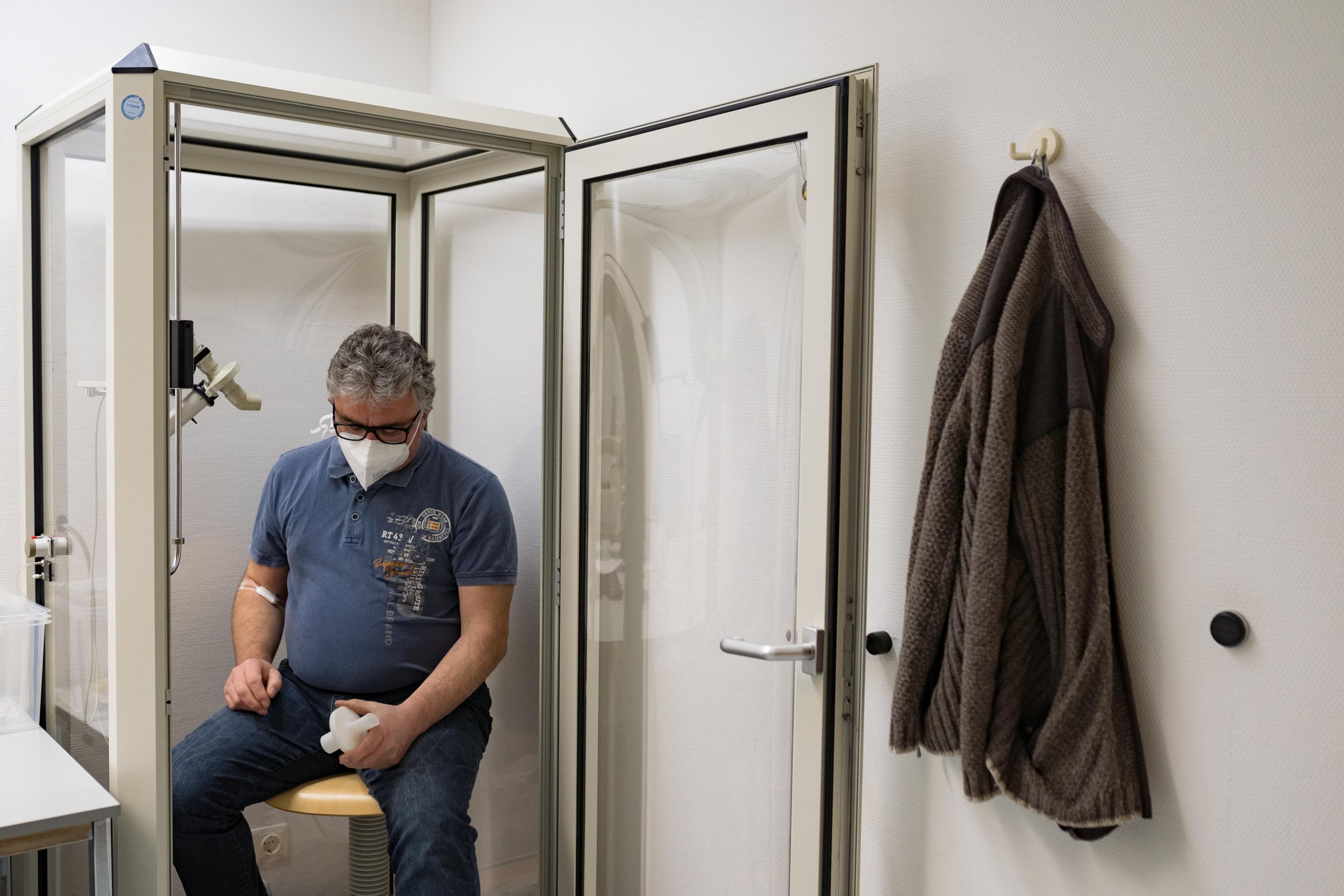 Ein älterer Mann sitzt zusammengesunken in einer Glaskabinen, in der Lungenfunktionstests gemacht werden. Er blickt auf einen Plastikaufsatz in seiner Hand, durch den er in der Kabine durchpusten muss.