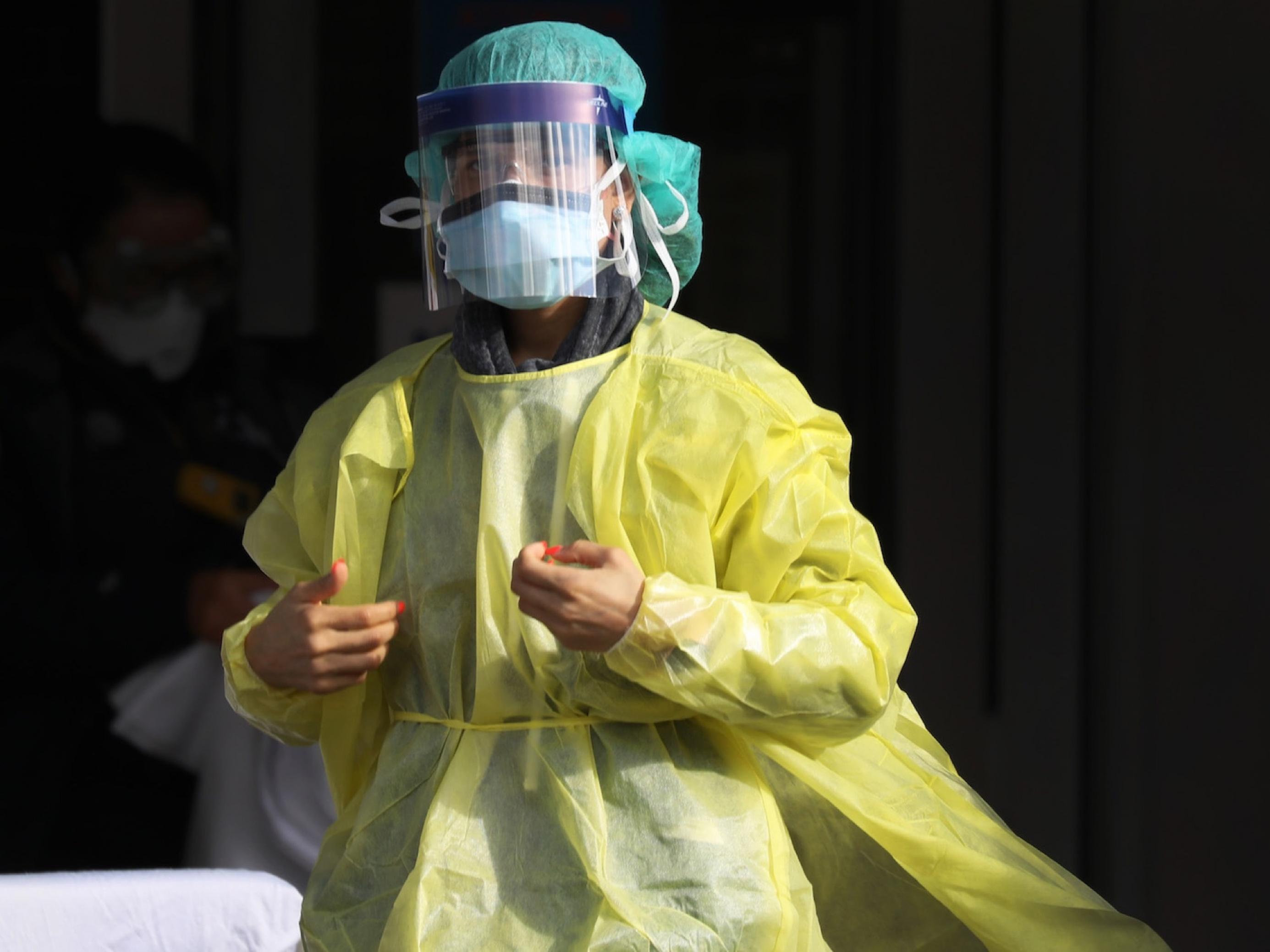 Das Bild zeigt eine Frau, die komplett in medizinische Schutzkleidung aus Plastik gehüllt ist. Man sieht ihre rot lackierten Fingernägel, aber ansonsten wenig. Sie steht vor dem Krankenhaus.
