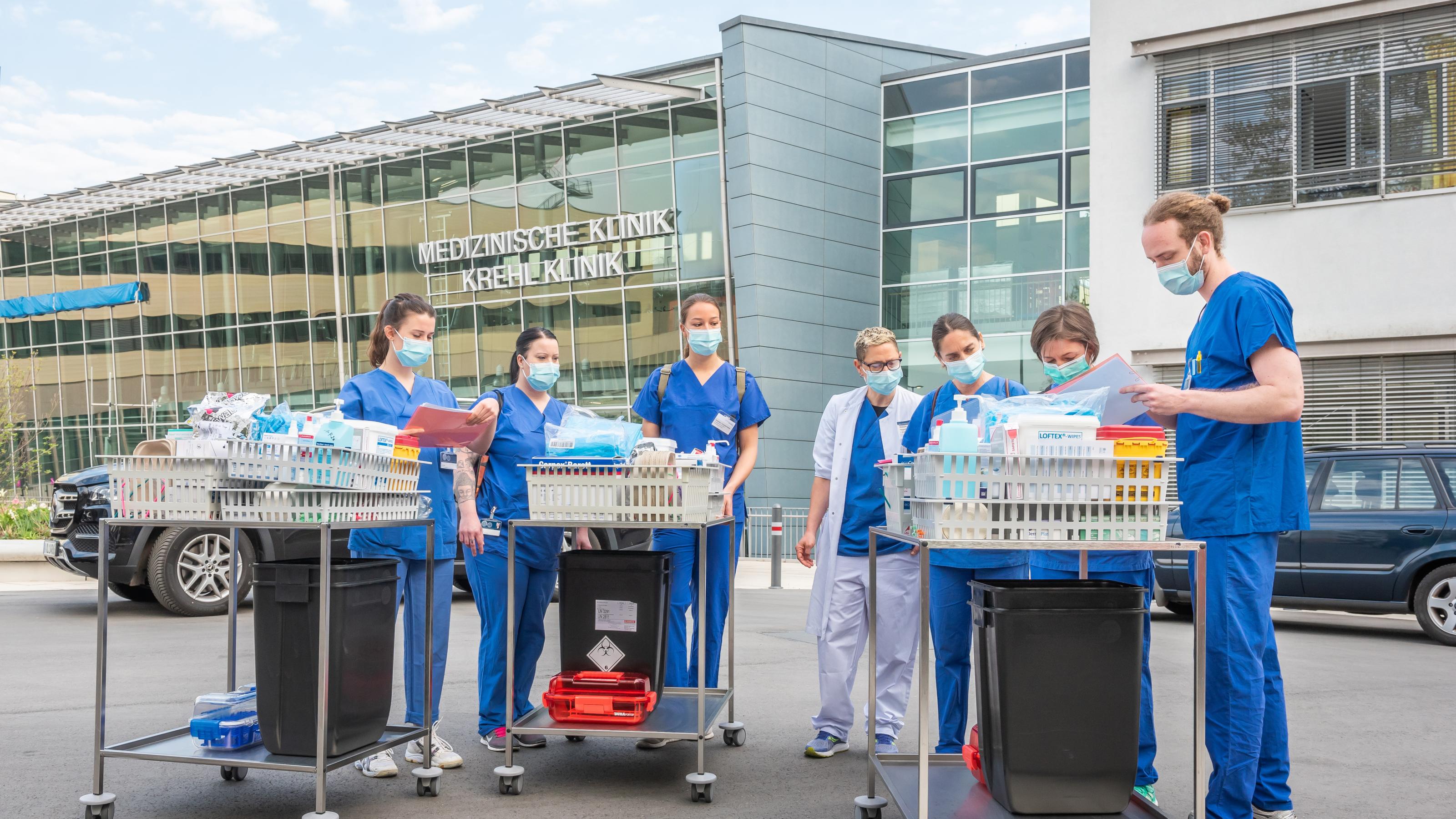 Medizinstudierende in blauer Arbeitskleidung und Mund-Nasenschutz bereiten sich auf eine Fahrt zu SARS-CoV-Infizierten vor (Coronataxi). Sie stehen vor einem Gebäude der Universitätsklinik Heidelberg