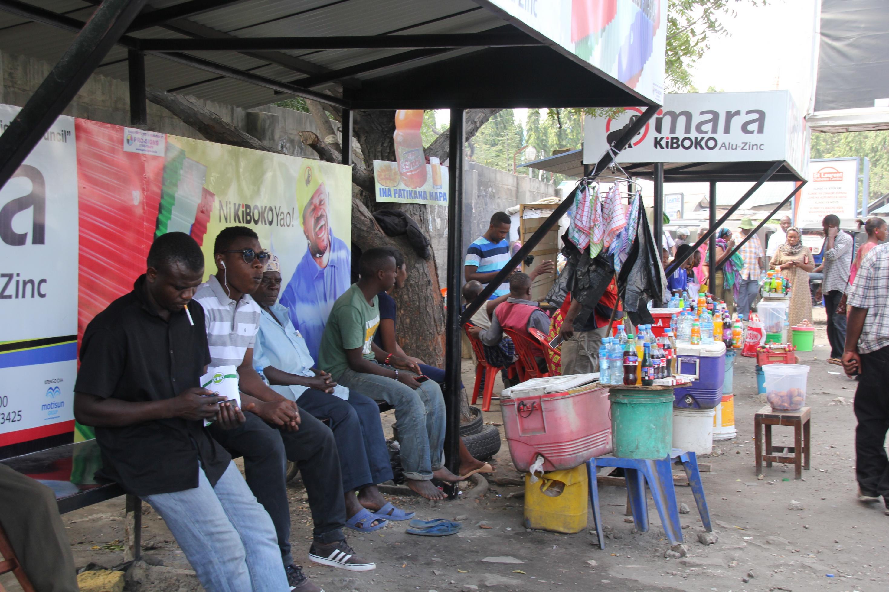 An einer Straße in der tansanischen Metropole Dar es Salaam warten viele Menschen an einer Bushaltestelle. Daneben haben Händlerinnen und Händler ihre Waren aufgebaut, darunter Limoflaschen, Tücher, Eimer mit Fettgebackenem.