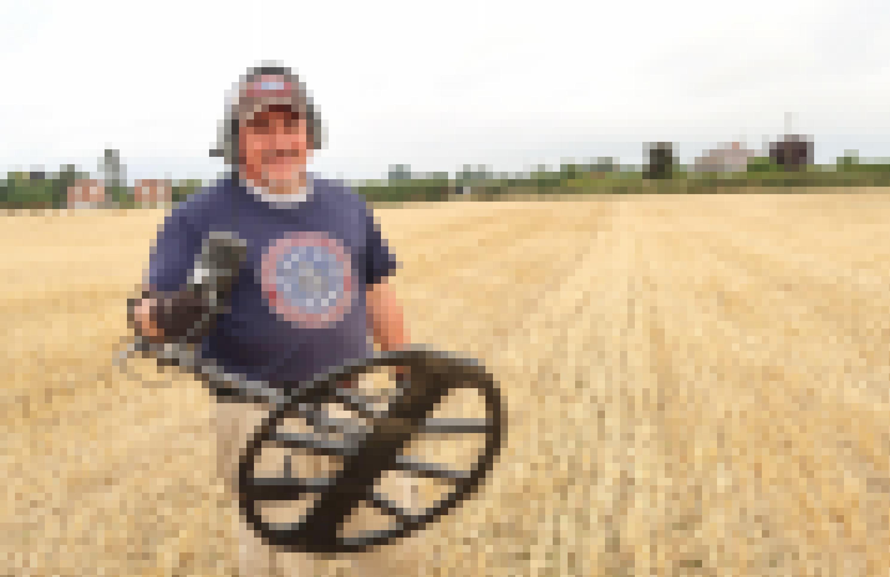 Ein Mann hält das vordere Ende eines Metalldetektors in die Kamera. Er steht mitten auf einem großen Feld.