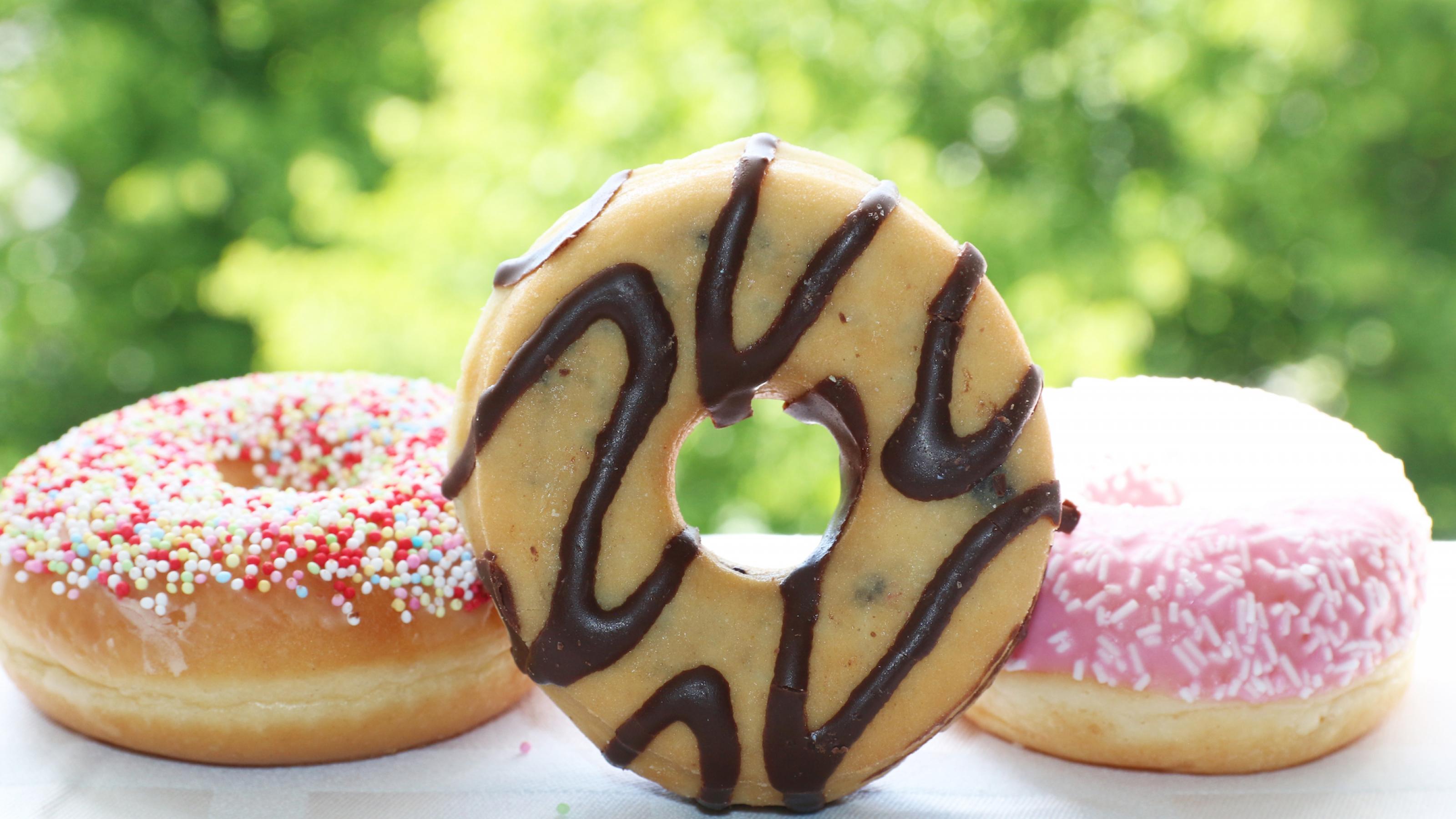Drei Donuts liegen auf einen weißen Tuch vor einem offenen Fenster, der mittlere steht so auf der Seite, dass man durch das Loch blicken kann. Der linke ist mit Zuckerperlen verziert, der mittlere mit Schleifen von Schokoguss, der rechte mit rosa Zuckerguss. – Hefeteig, Loch in die Mitte, frittieren und verzieren – Doughnut oder Donut heißt die Kalorienbombe.