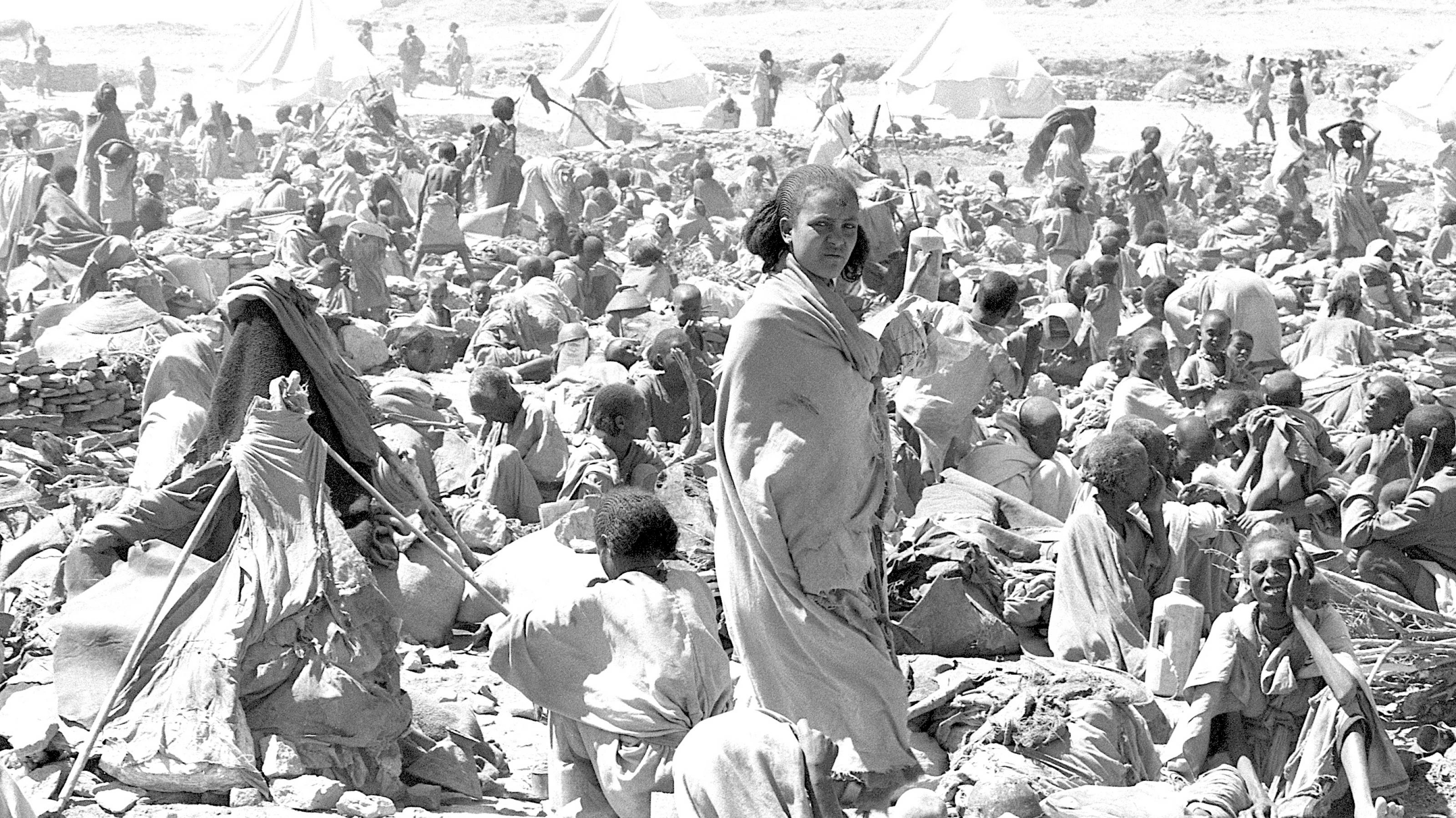 In einer Wüstengegend warten Dutzende Menschen auf Hilfe. In der Mitte des Bildes steht eine junge Frau und blickt ernst in die Kamera. Ein Flüchtlingslager während der Hungerkrise 1984 in Äthiopien. Foto in Schwarz-Weiß in Sepia-Tönen.