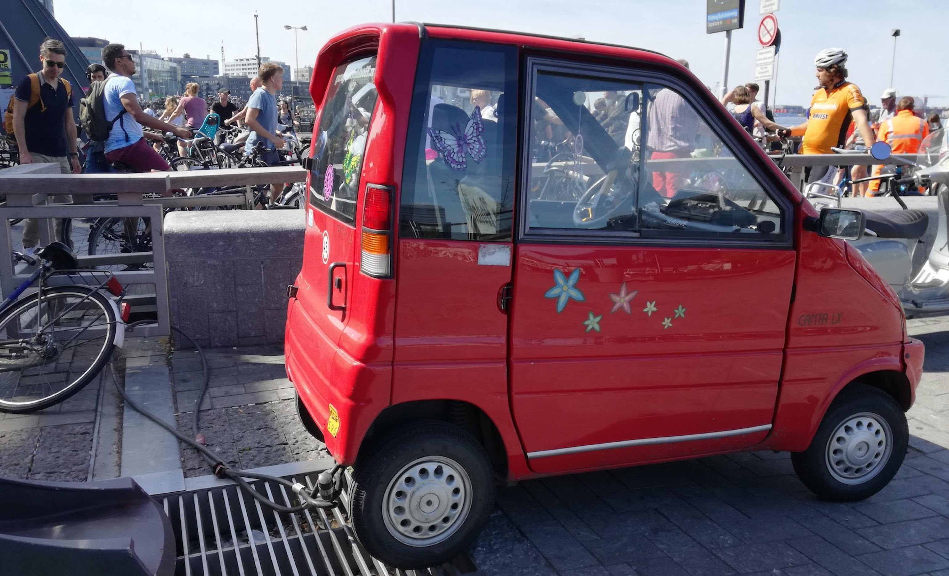 Ein kleines rotes E-Auto steht an einer Ladestation. Im Hintergrund sind Menschen zu sehen.