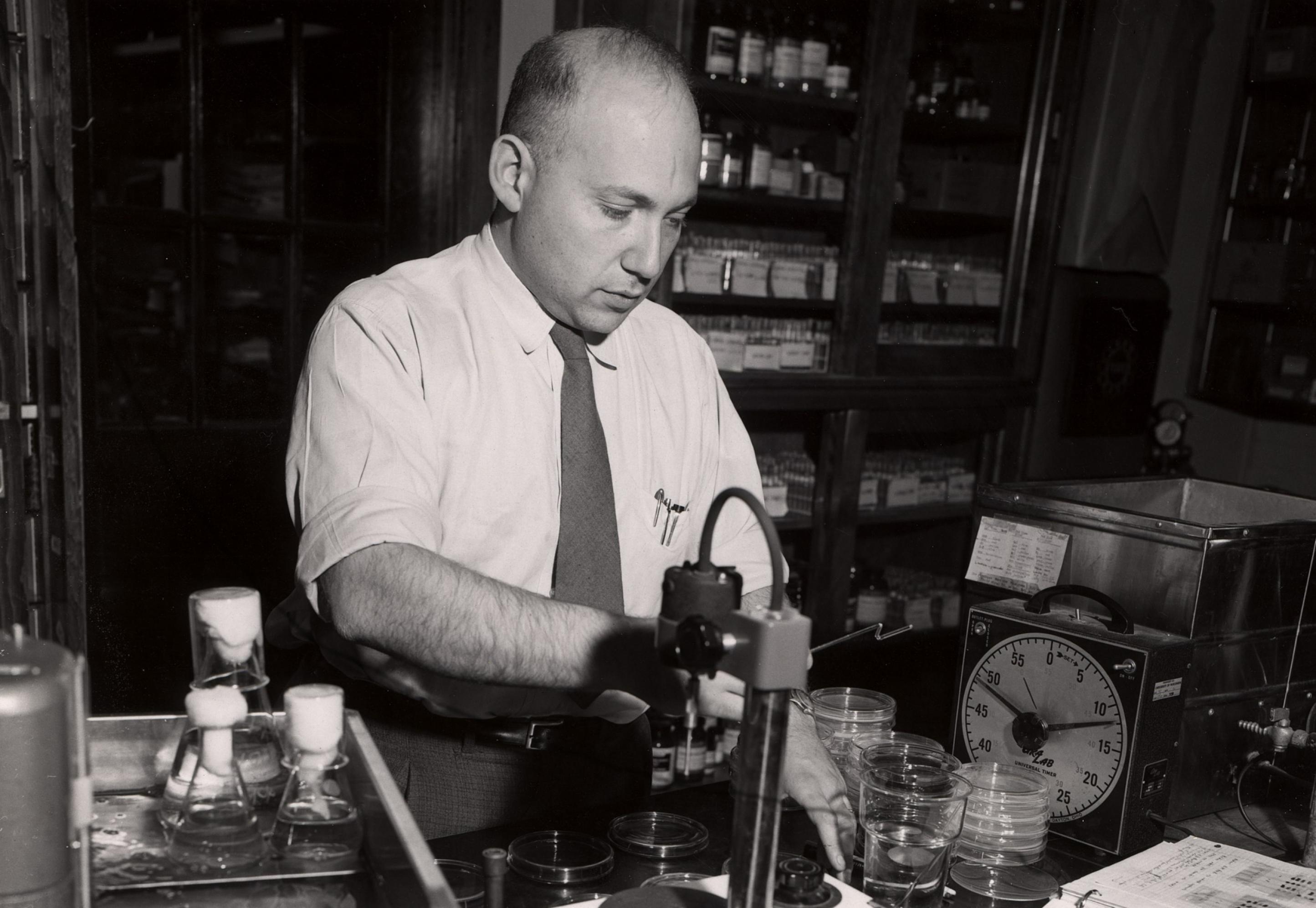 Schwarz-Weiß-Foto des Nobelpreisträgers Joshua Lederberg, der in Hemd und Krawatte in seinem Labor mit Bakterienkulturen arbeitet.