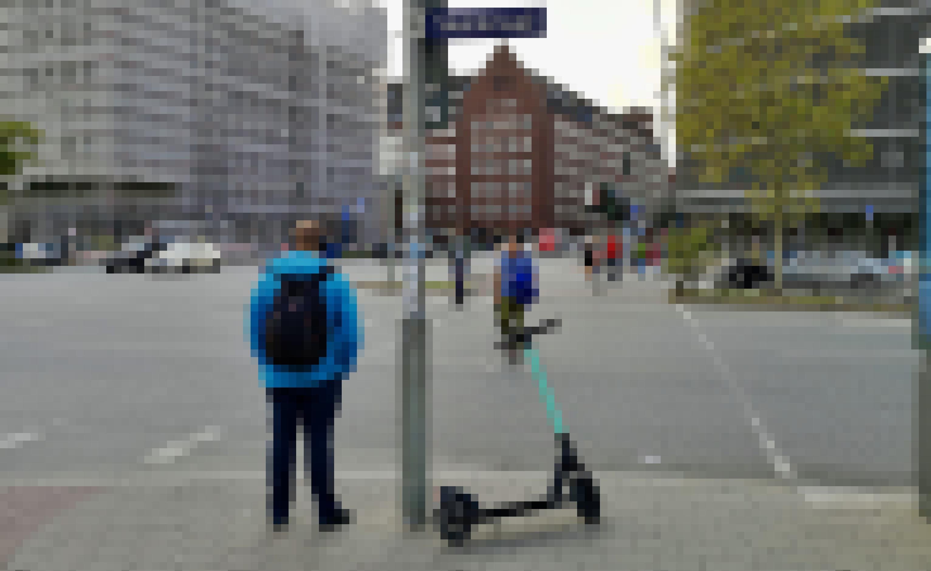 Ein E-Kick-Scooter wurde auf einer Verkehrsinsel abgestellt. Links wartet ein Mann auf das Grünsignal für Fußgänger.