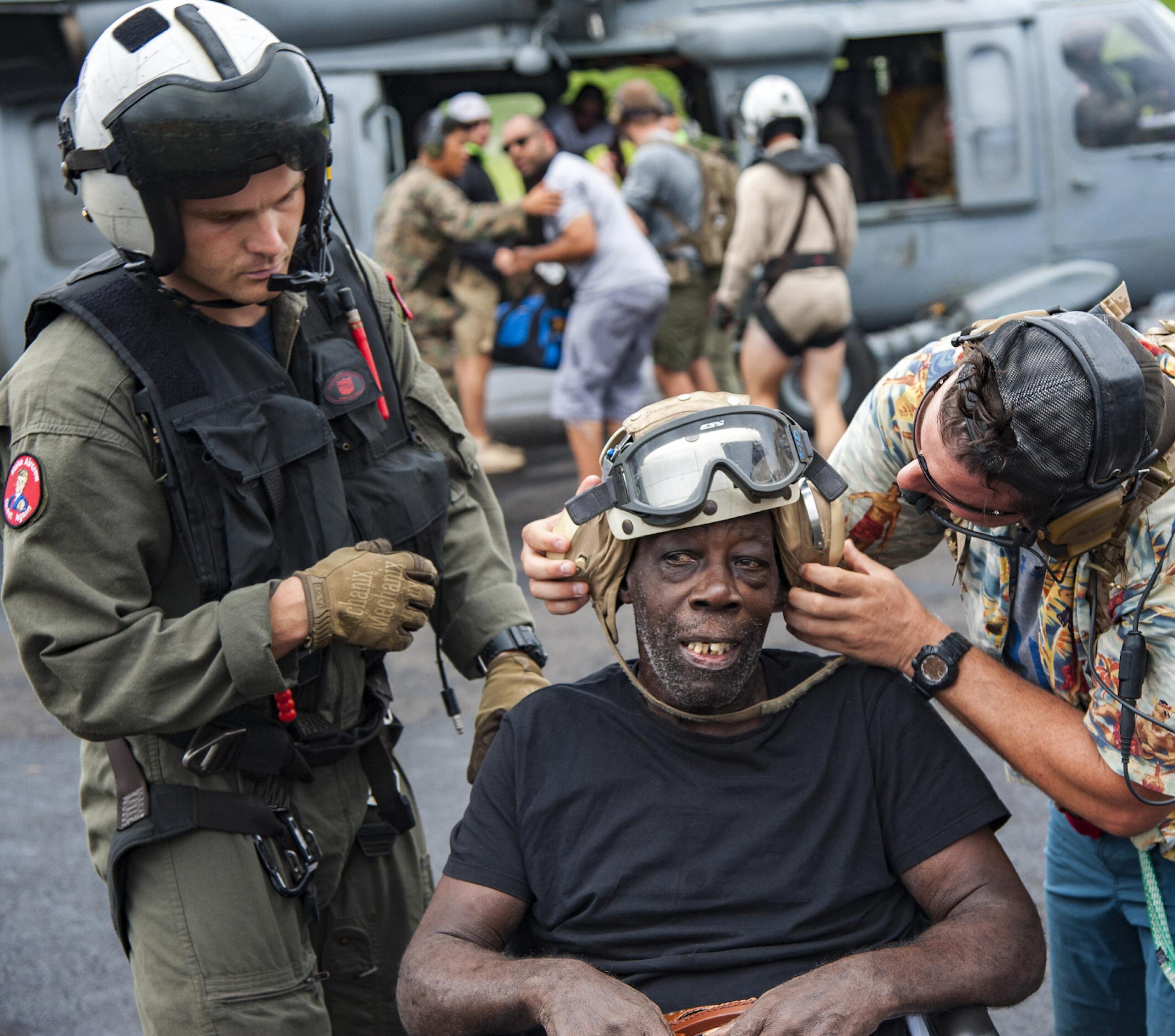 Ein Soldat und ein zweiter Helfer setzen einem alten Mann eine Fliegerkappe mit Schutzbrille und Kopfhörer auf. Als Hurrikan „Maria“ im September 2017 die Karibikinsel Dominica platt gemacht hatte – anders kann man es nicht sagen – blieb den Überlebenden nur noch die Evakuierung. Auch die US-Marine half mit ihren Hubschraubern.