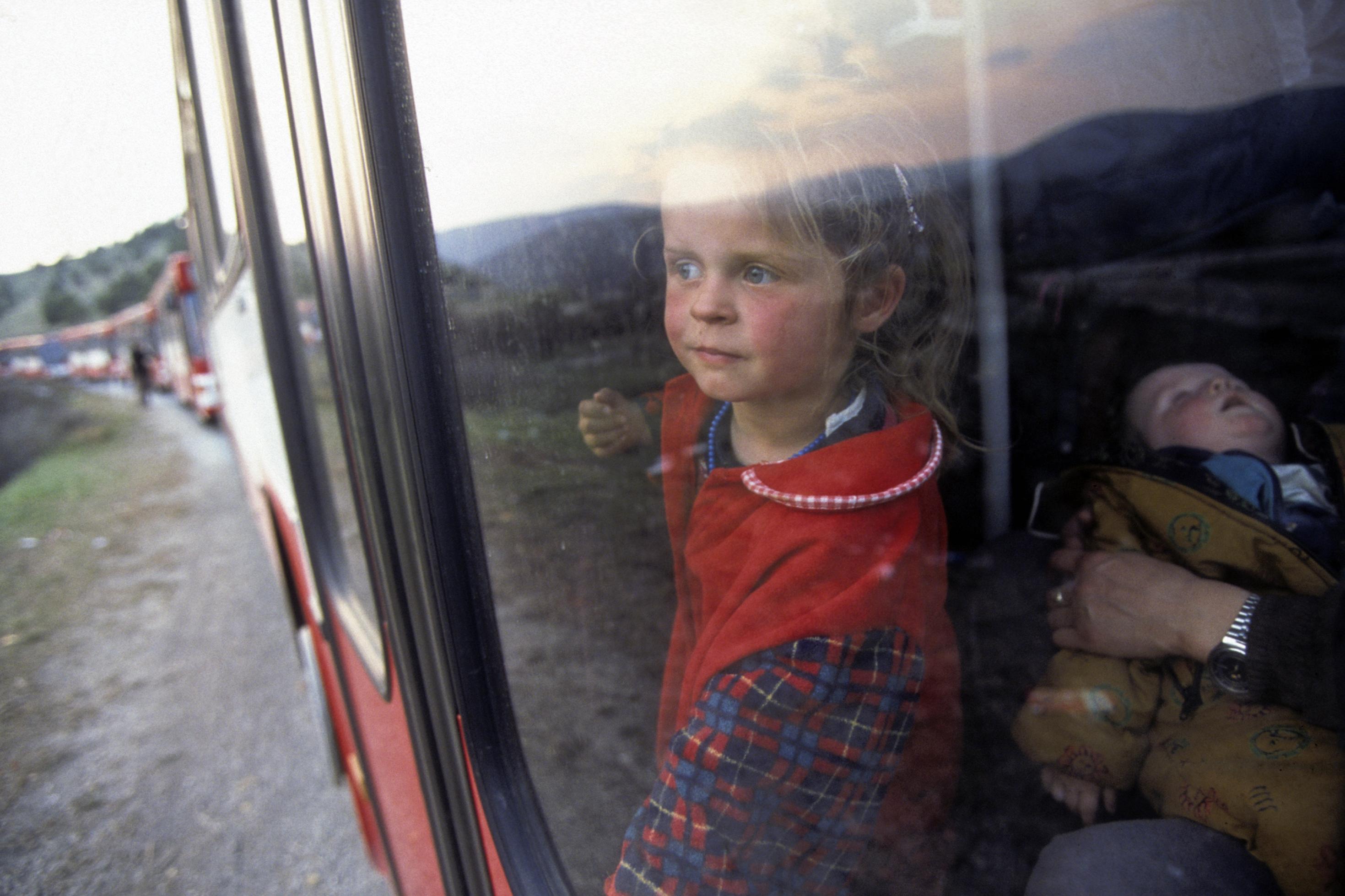 Eine Mädchen steht an der Glastür eines Busses und schaut hinaus. Neben ihr hält eine Frau ein Baby im Arm. Im Hintergrund reihen sich viele weitere solche Busse. Die Karawane bringt im Jahr 1999 Kosovo-Flüchtlinge aus der Gefahrenzone; die Gruppe soll in Deutschland Schutz finden.